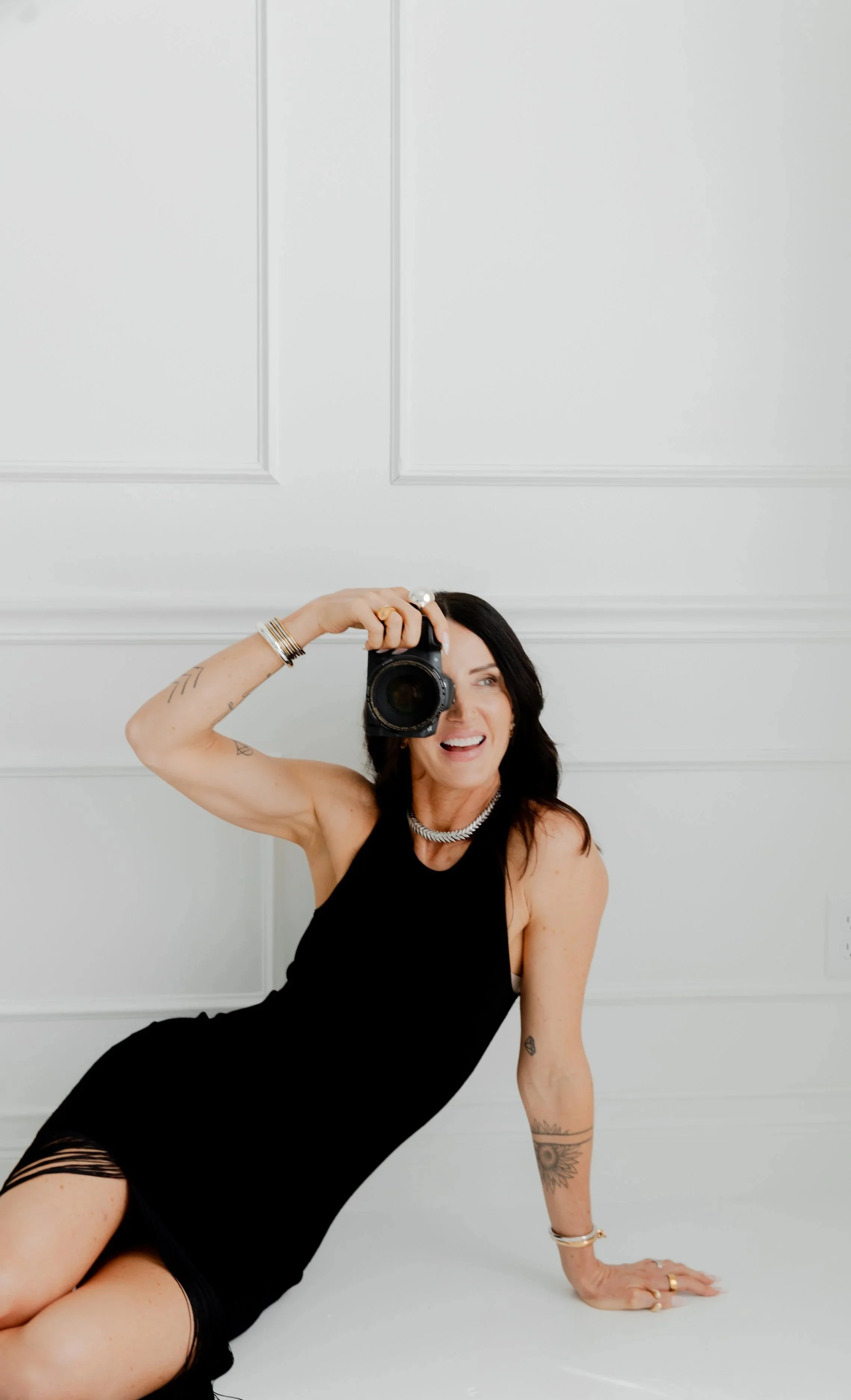 Woman in black dress taking a selfie with a camera, sitting on the floor in front of a white paneled wall.