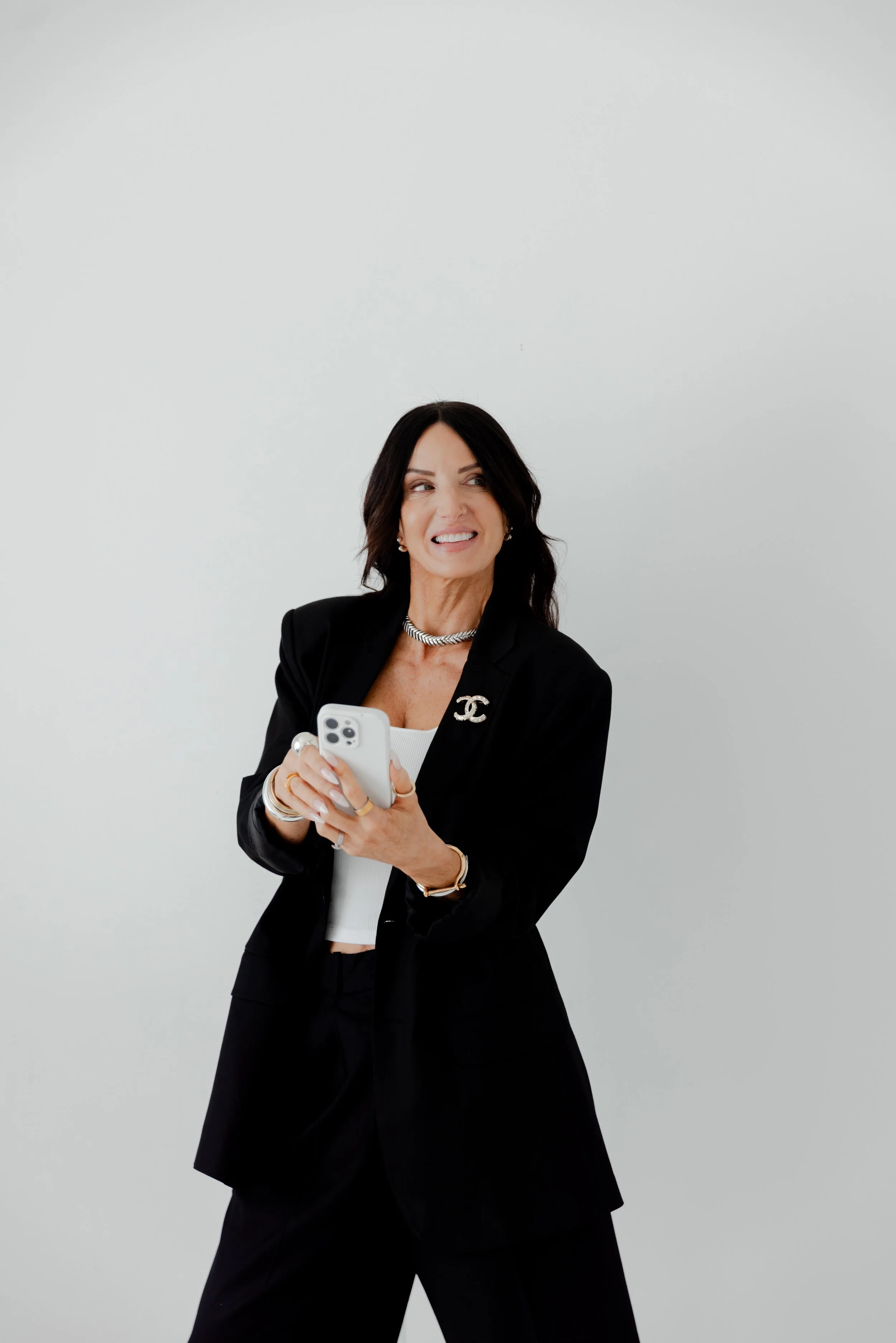A woman with dark hair wearing a black blazer and pants, holding a smartphone, smiling with a slightly mischievous expression against a plain white wall.