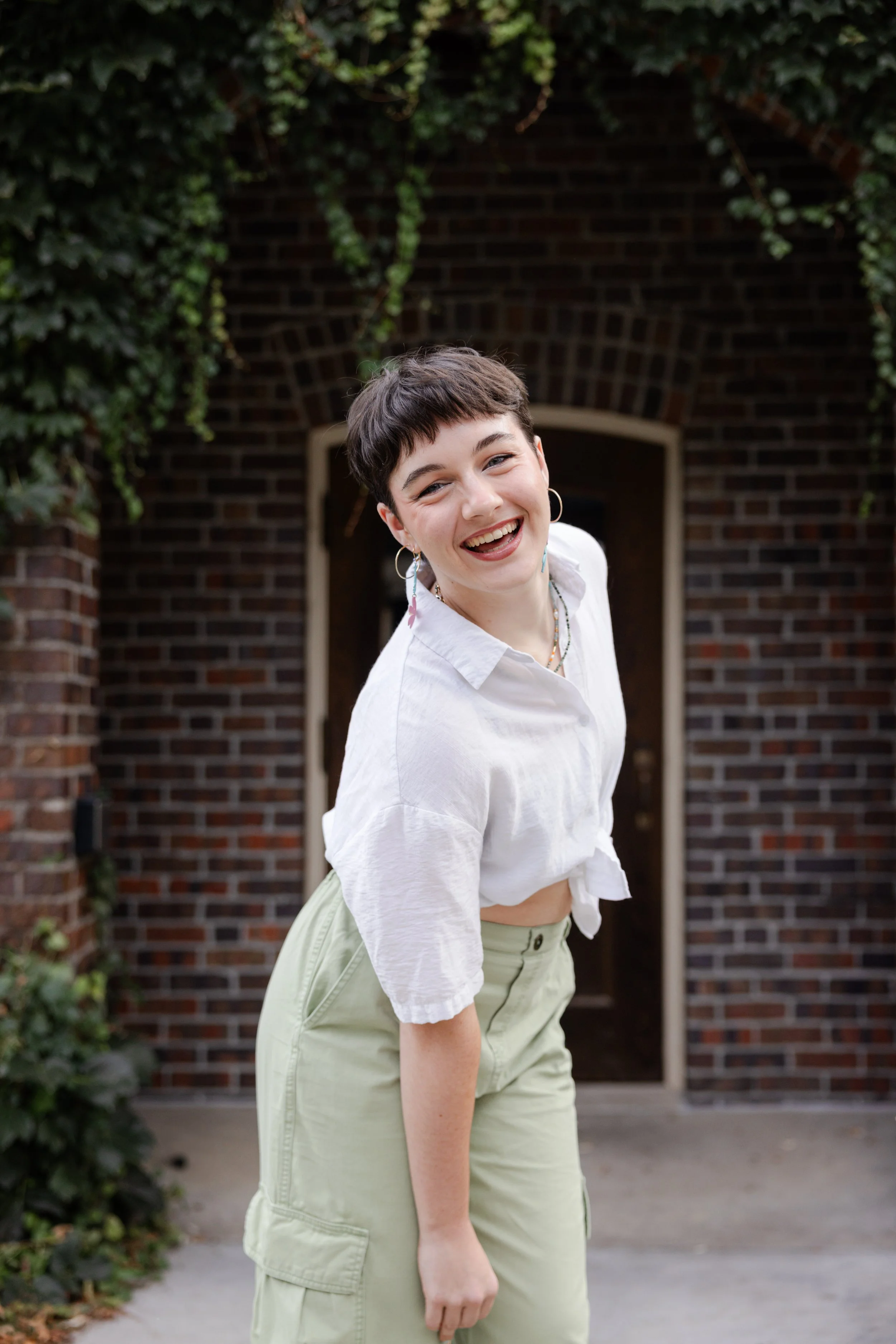 A young woman with short dark hair, wearing a white crop top, high-waisted light green cargo pants, hoop earrings, and a colorful beaded necklace, smiling and tilting her head to the side outdoors in front of a brick and ivy wall.