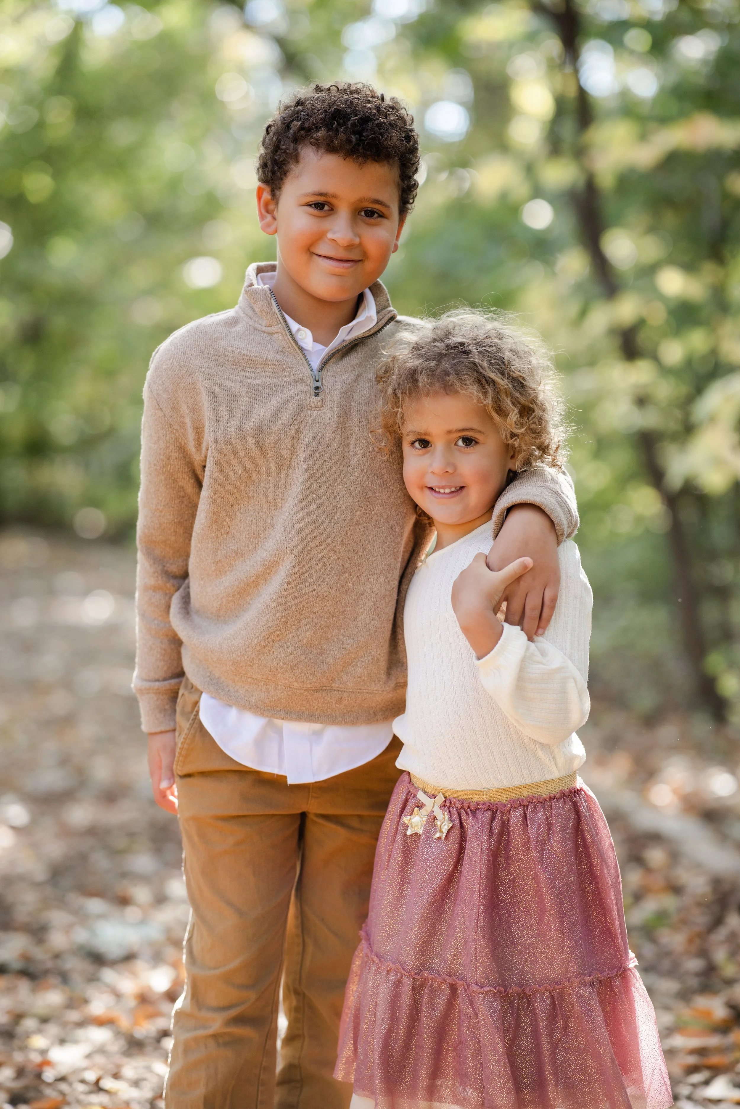Two children, a boy and a girl, standing outdoors in a park surrounded by trees with sunlight filtering through, smiling at the camera.