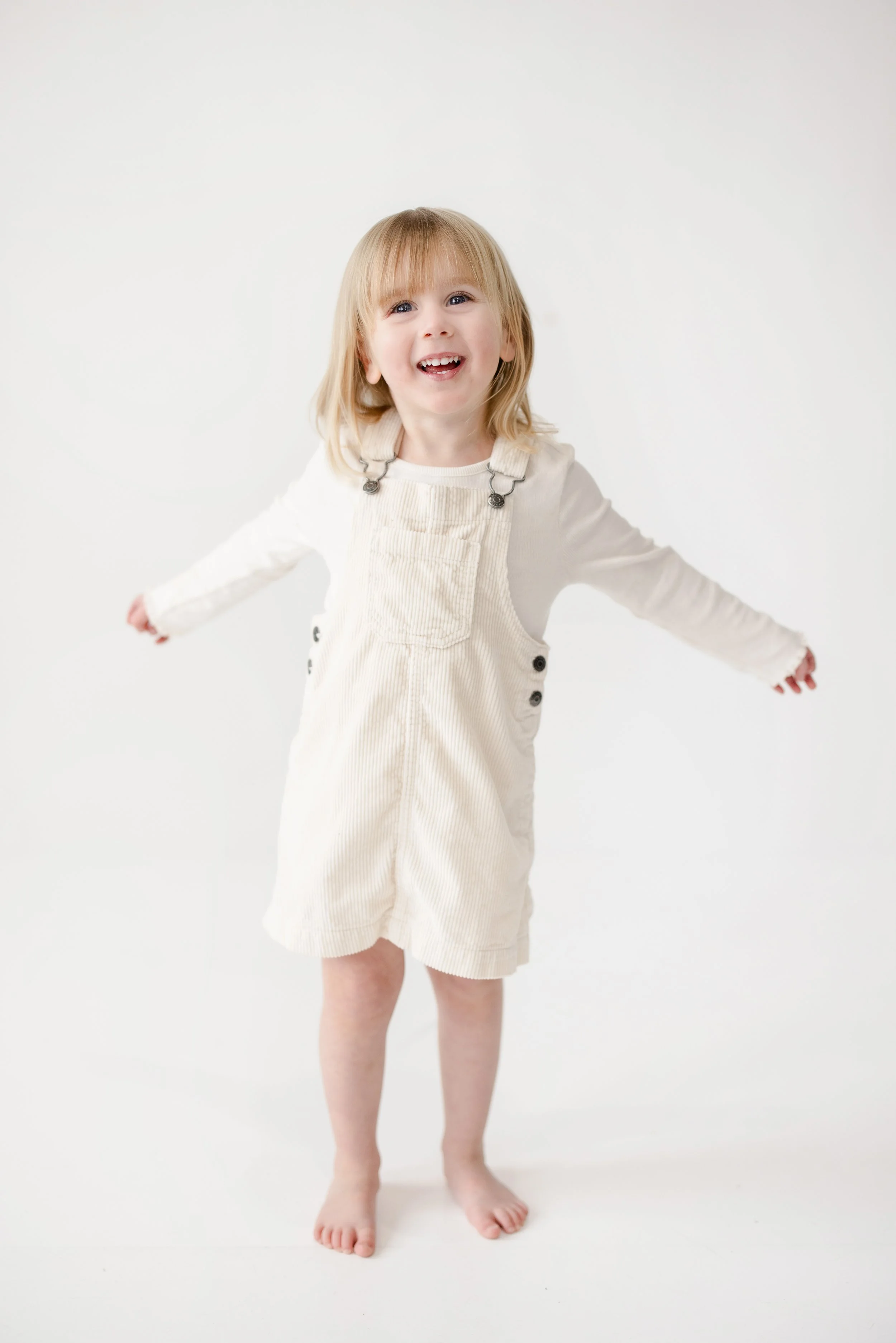 A young girl with blond hair and a big smile standing against a white background, wearing a cream-colored romper and barefoot.