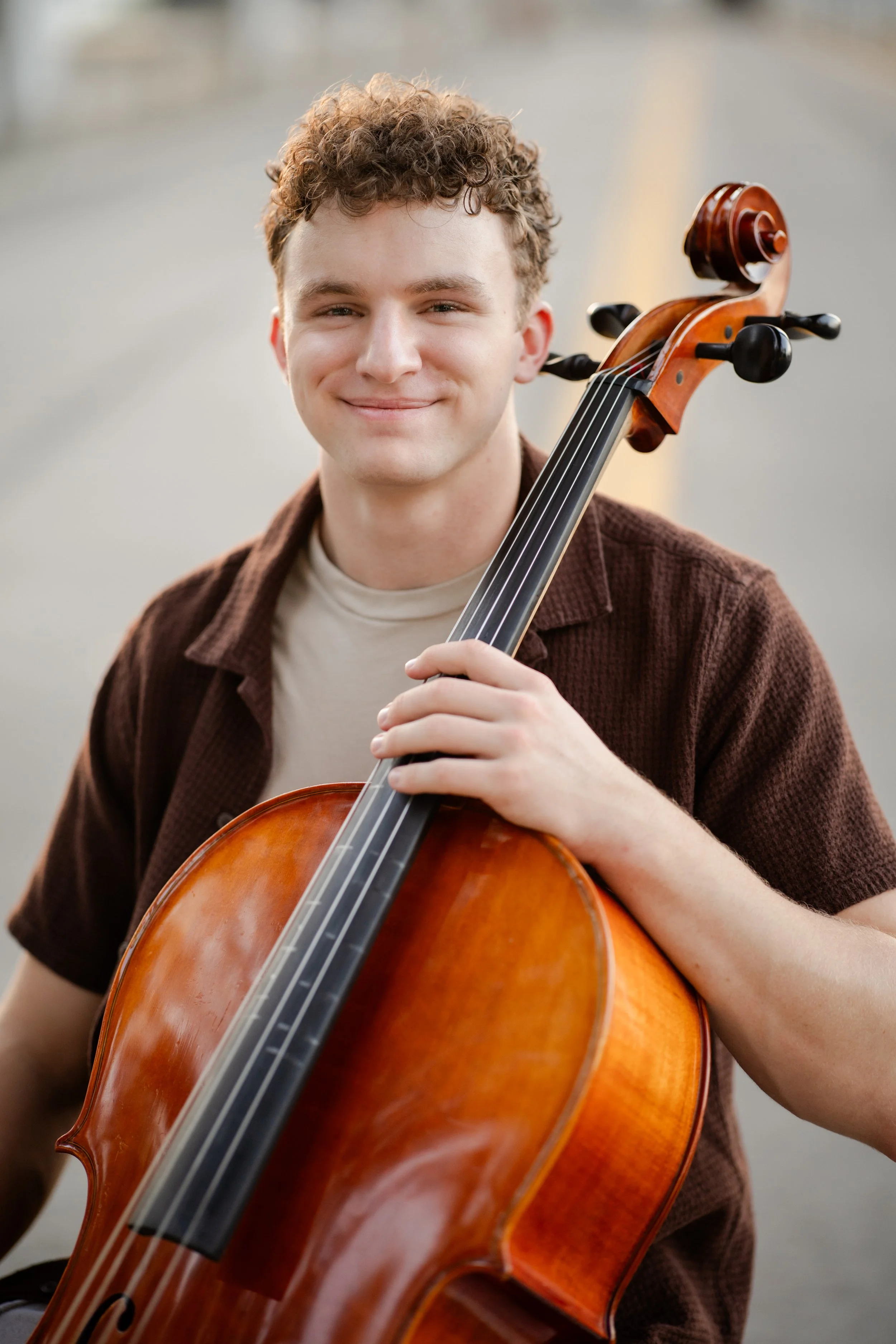 A young man with curly hair smiling while holding a cello.