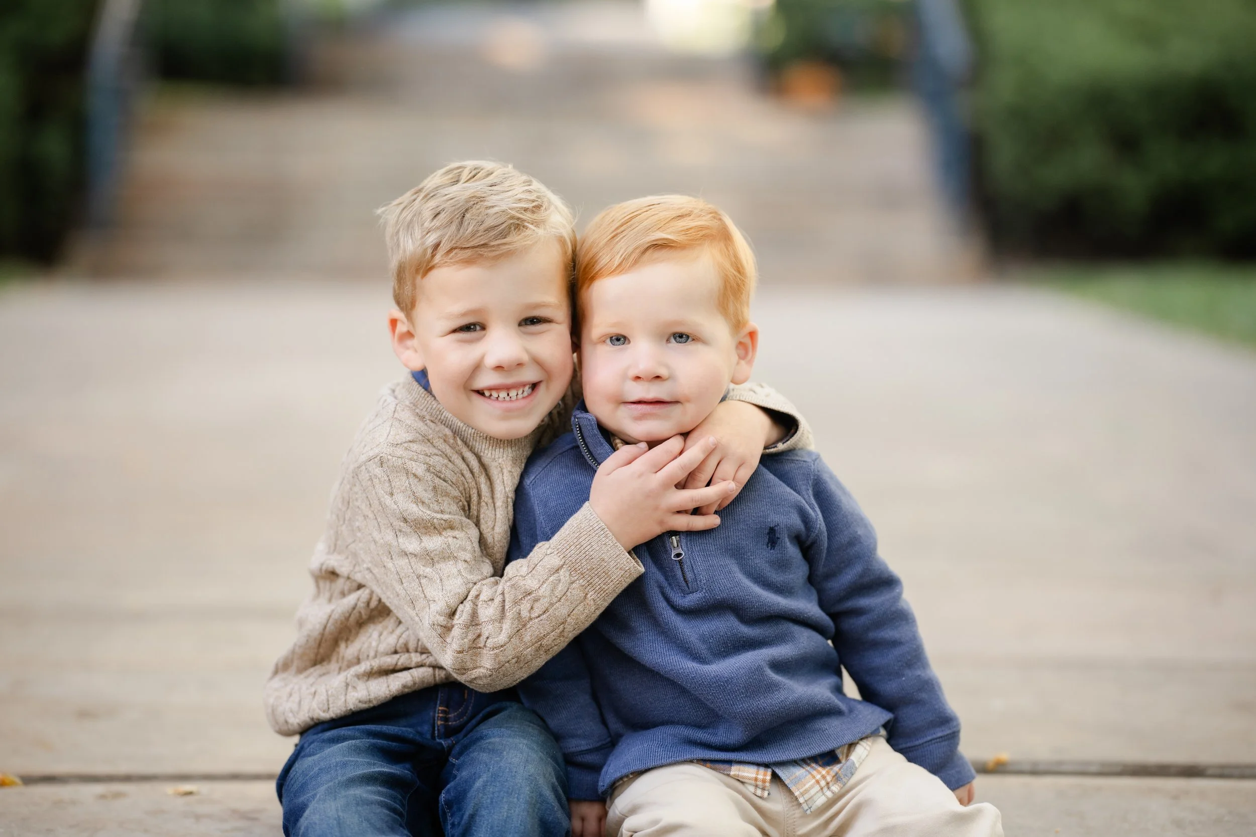 Two young boys, one with blonde hair and the other with red hair, sitting together on a playground. The blonde boy is smiling and hugging the red-haired boy, who has a neutral expression.