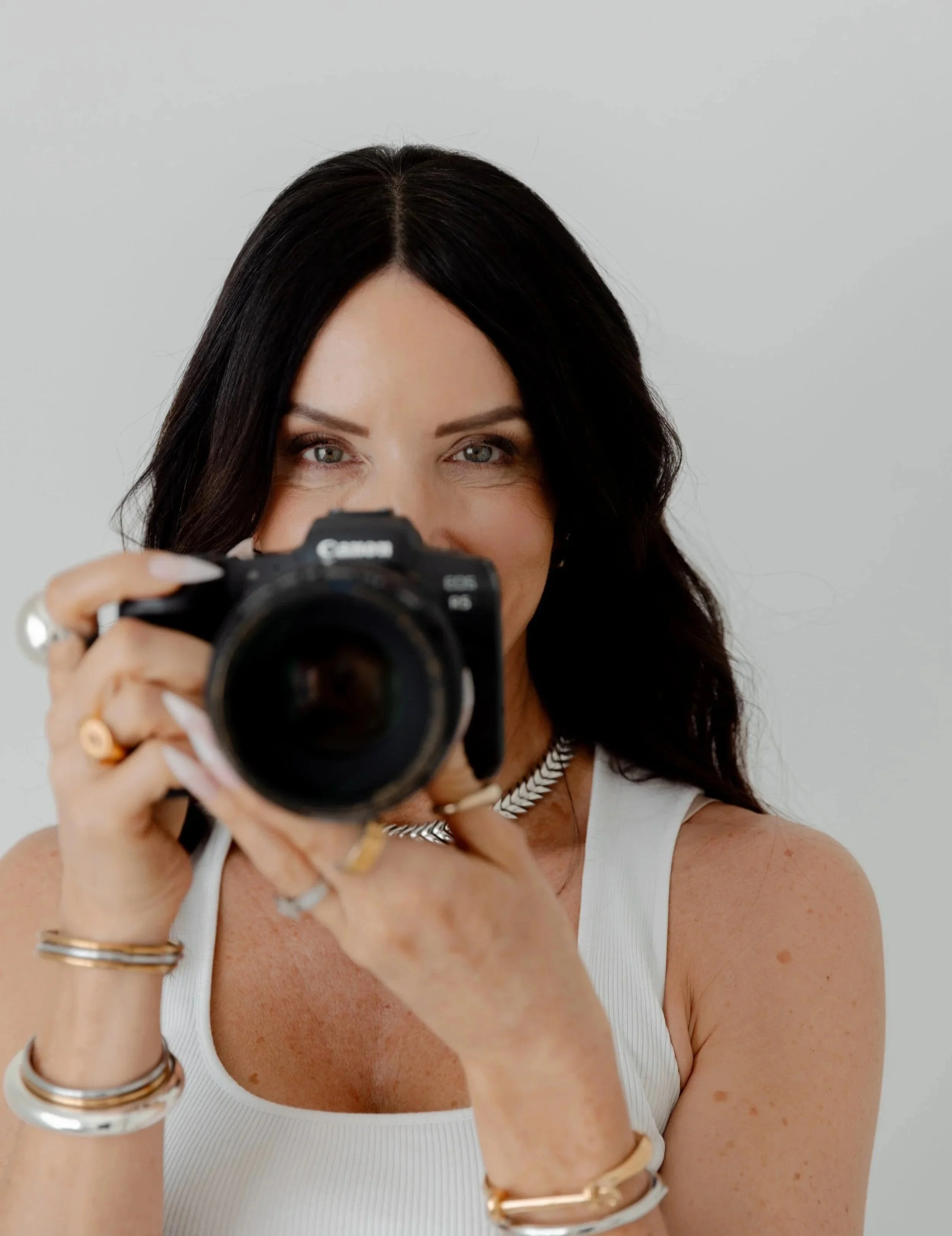 A woman with black hair and blue eyes taking a photo with a camera. She is wearing a white sleeveless top and jewelry, including rings, bracelets, and a necklace. The background is plain and light-colored.