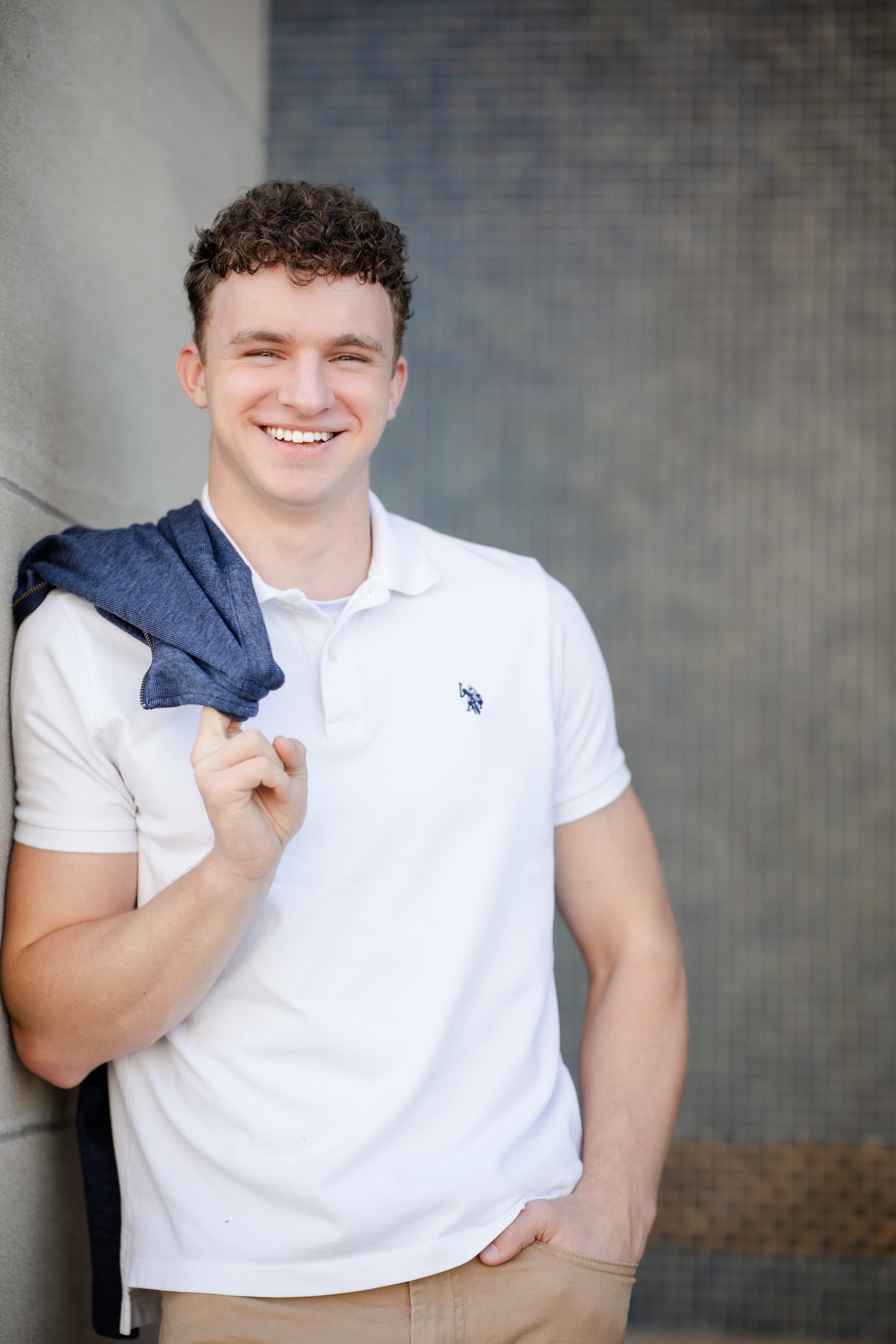 A young man with curly brown hair smiling and holding a navy blazer over his shoulder. He is leaning against a concrete wall, wearing a white polo shirt and beige pants, with a blurred background.