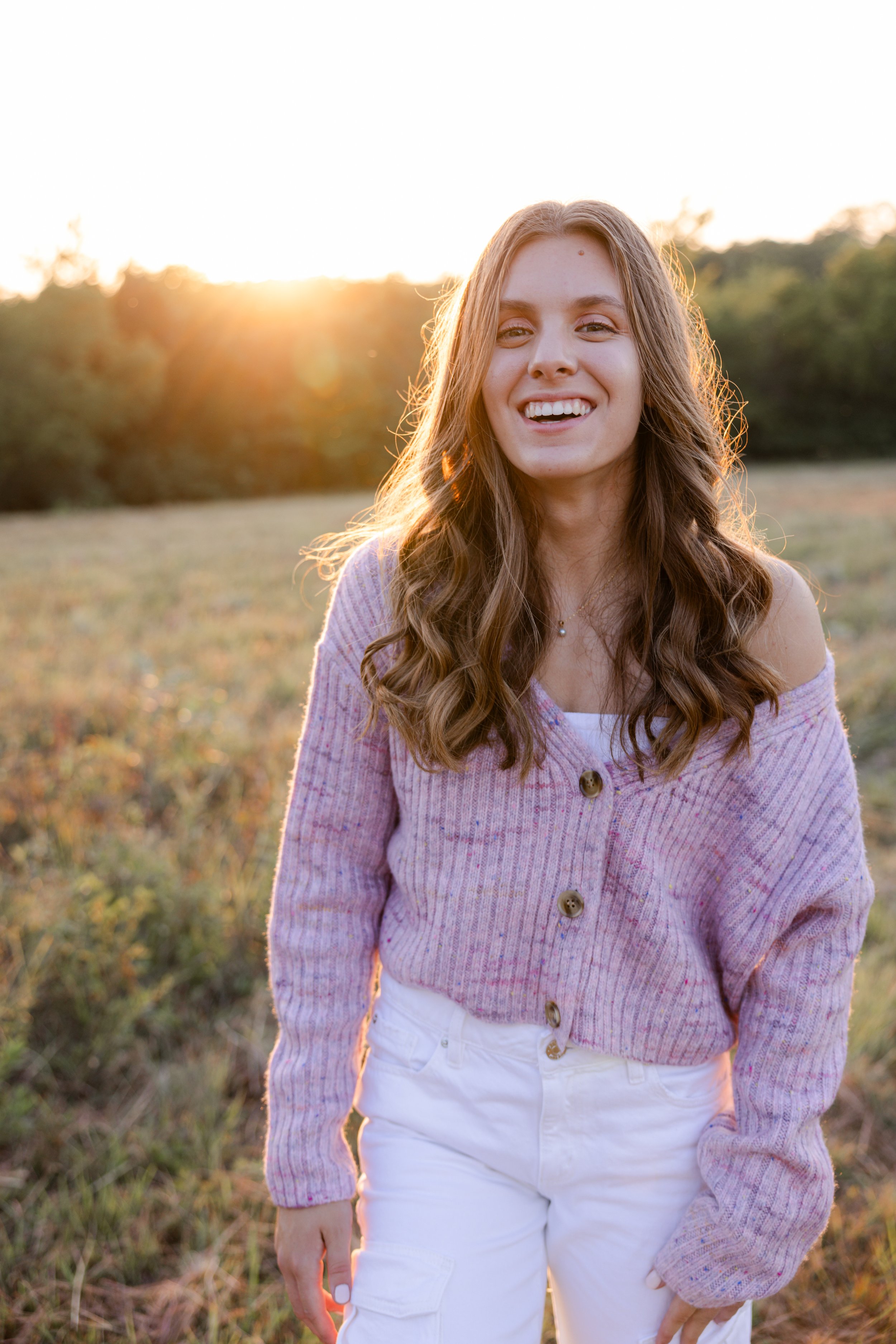 A young woman with long, wavy brown hair smiling outdoors at sunset, wearing a lavender cardigan and white pants in a grassy field with trees in the background.