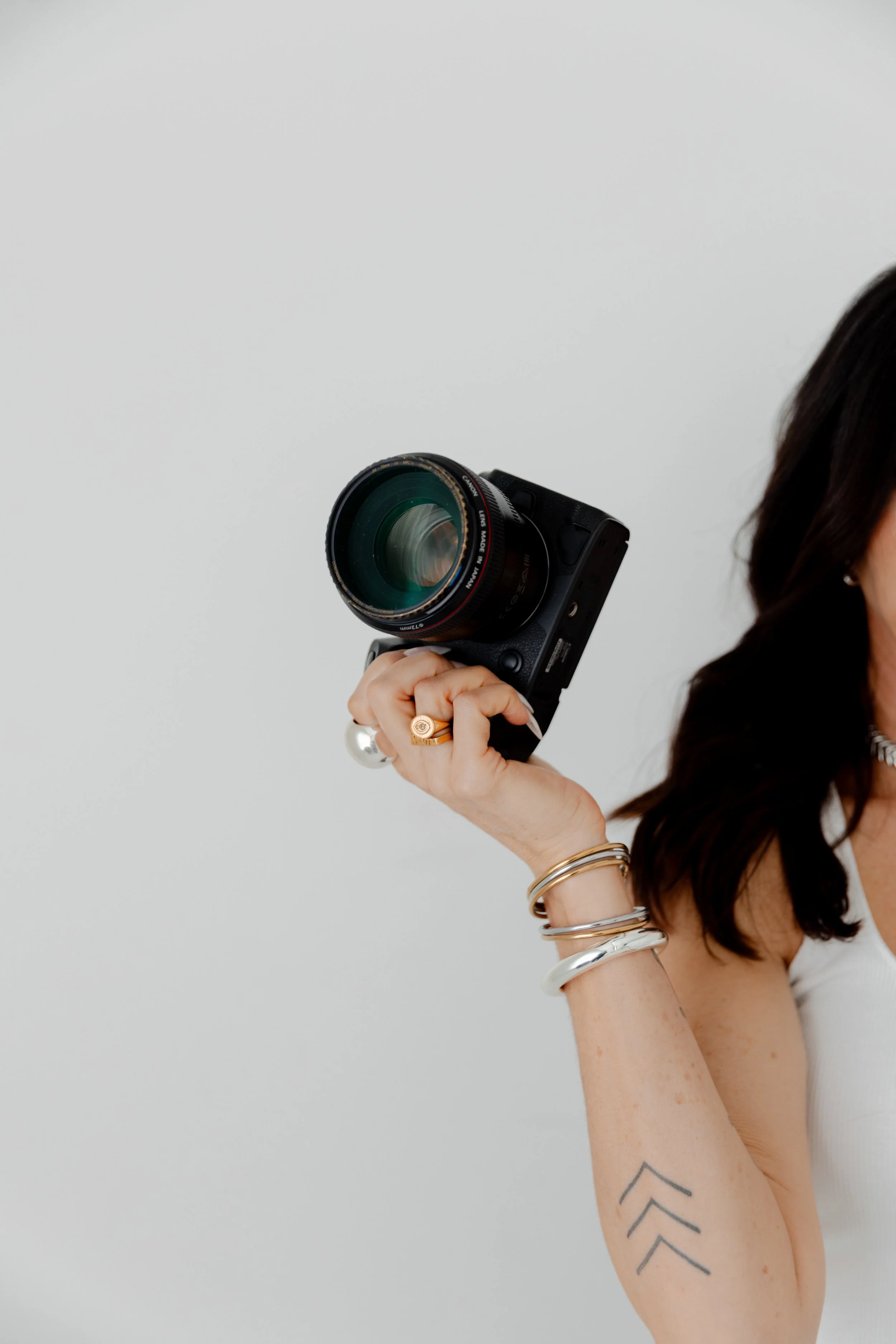 A woman with dark hair holding a professional camera with a large lens, wearing multiple bracelets and a ring, against a plain white background.