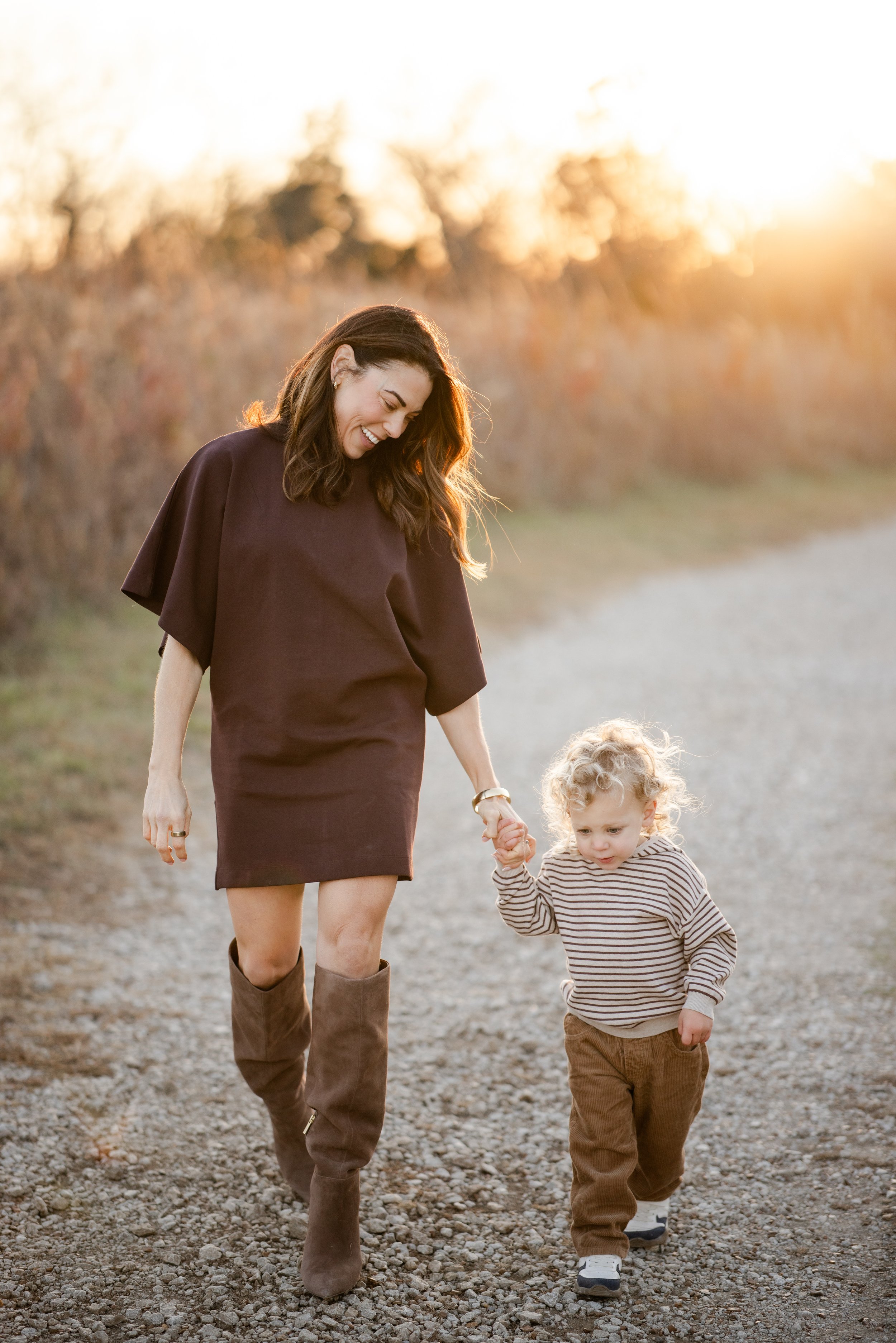 A woman and a young boy walking hand in hand on a gravel path during sunset, smiling and enjoying the moment together.