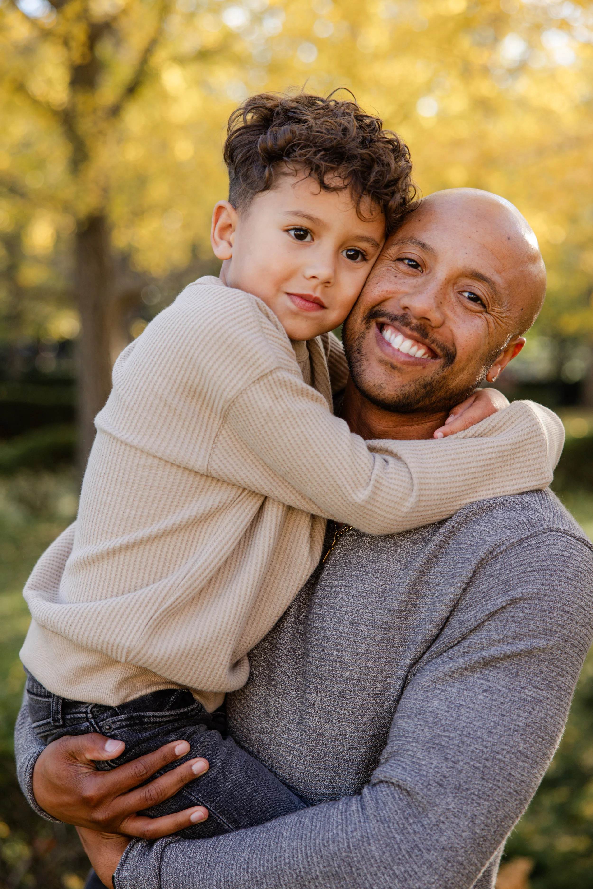 A happy man holding a young boy in an outdoor setting with autumn trees in the background.