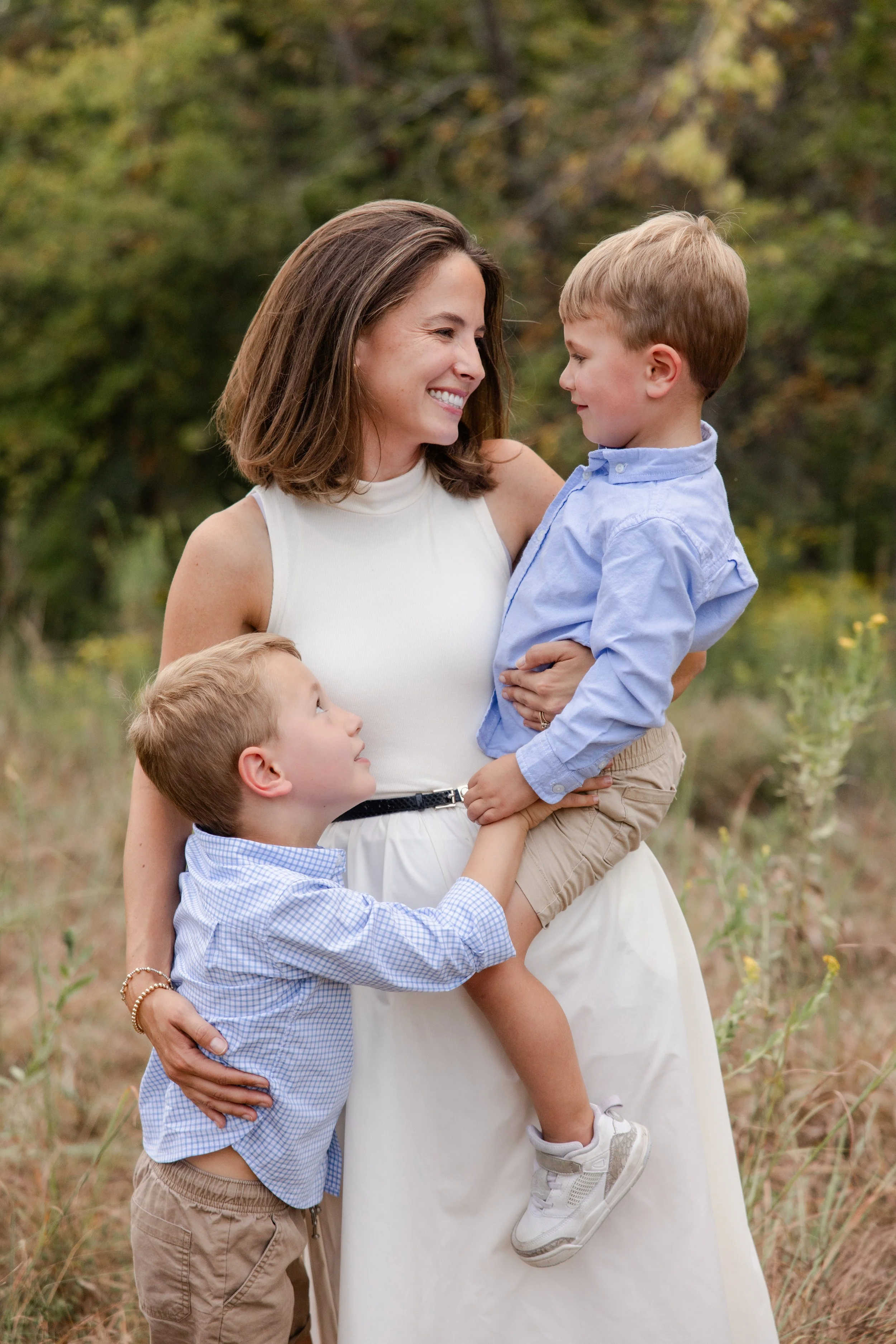 A woman smiling and holding a young boy in her arms while two other young boys stand beside her in an outdoor setting with trees and grass.