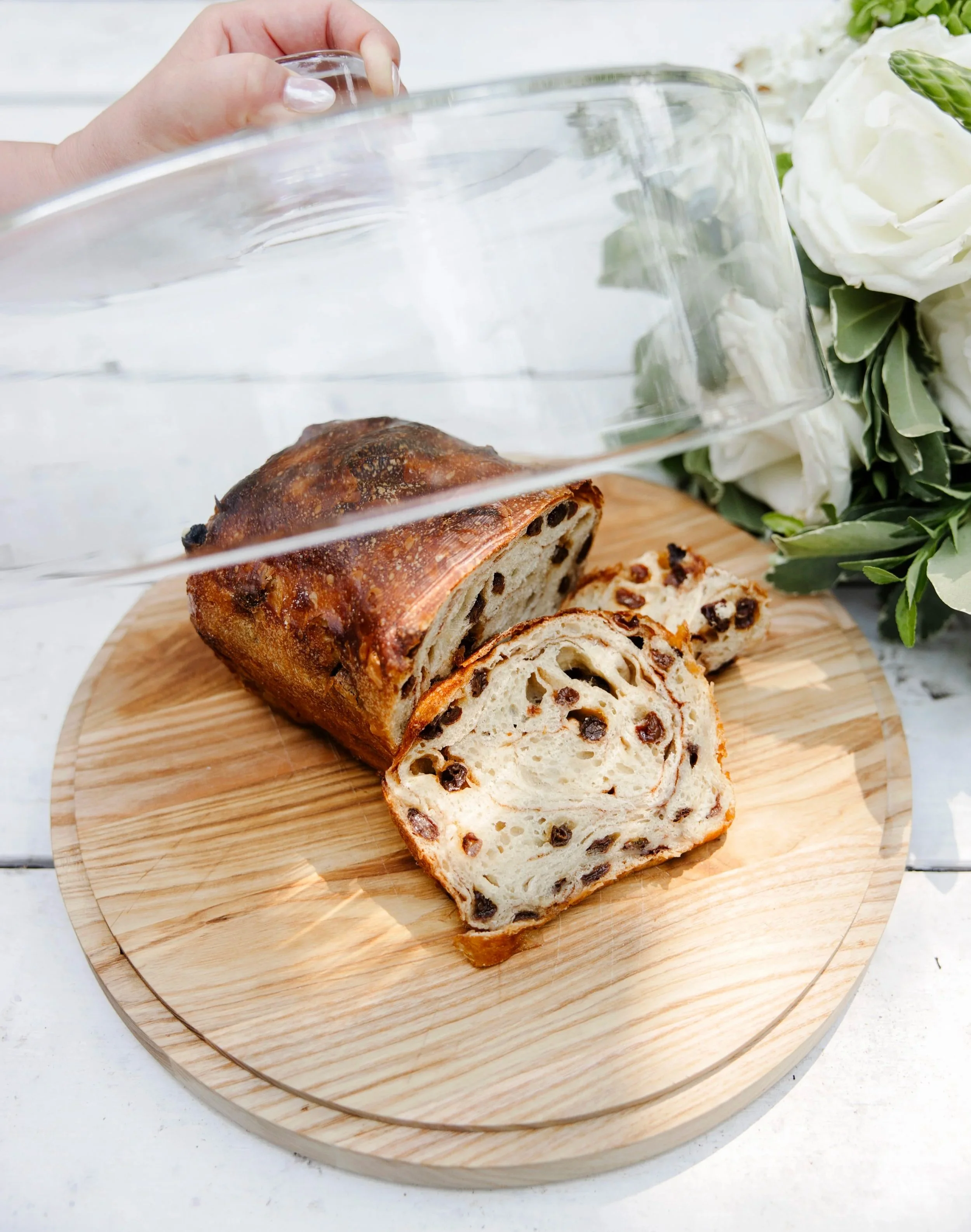 Sliced loaf of chocolate chip bread on a round wooden cutting board, with a bouquet of white flowers and greenery in the background.