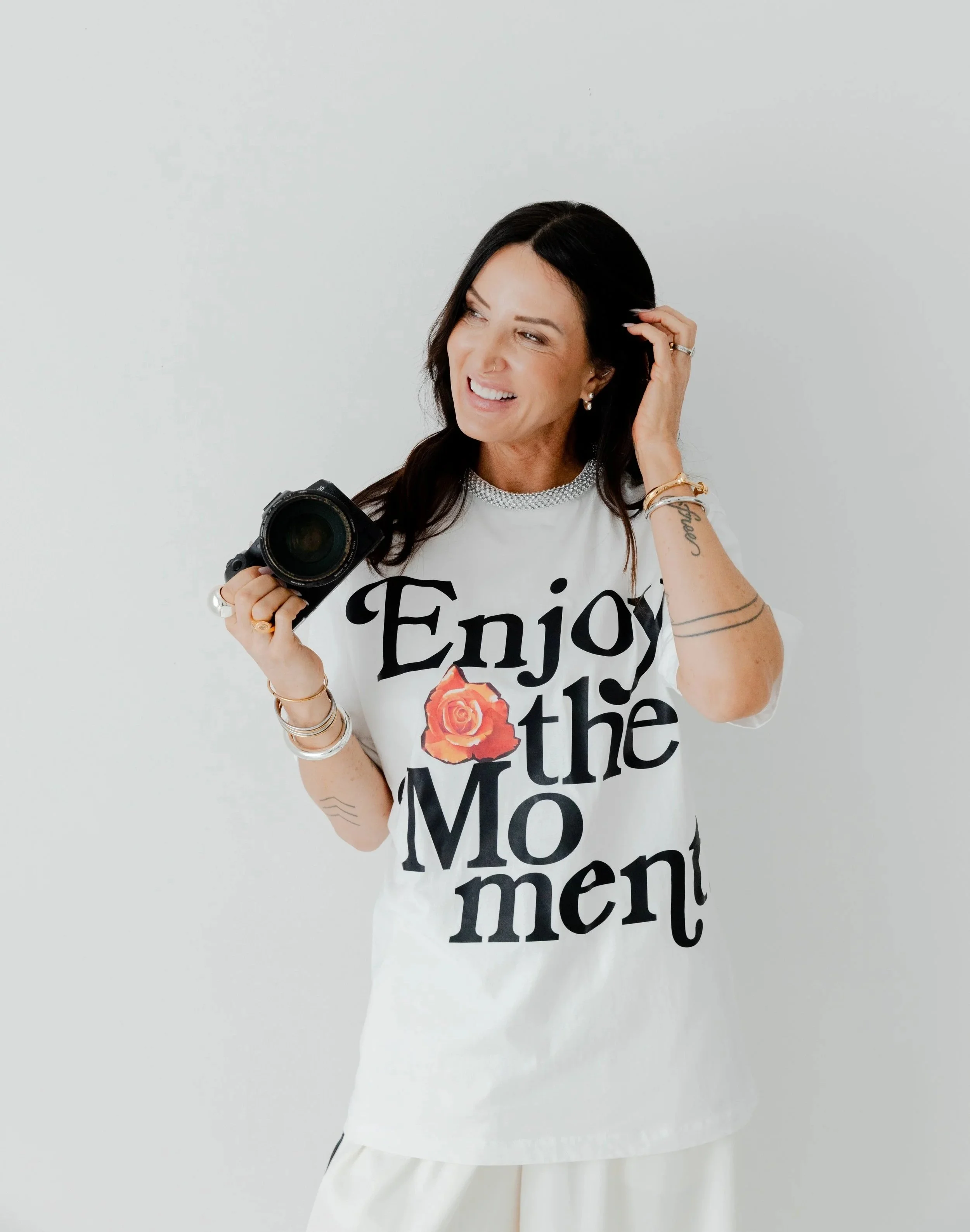 A woman with long dark hair and tattoos on her arm holding a camera, wearing a white t-shirt with bold black text that reads 'Enjoy the Moment' and a graphic of a pink rose.