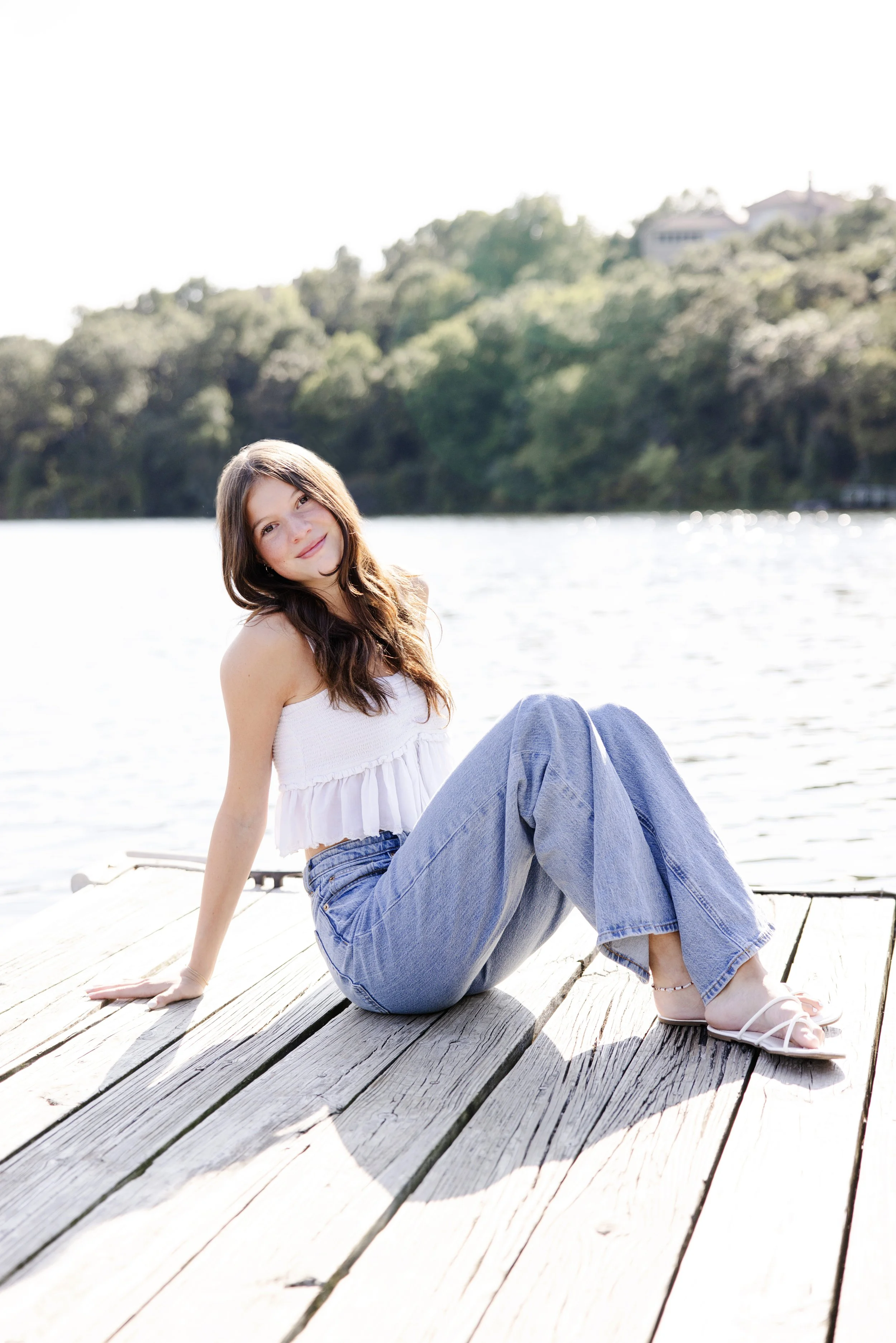 A young woman with long brown hair and fair skin sitting on a wooden dock by a body of water, smiling at the camera, wearing a white sleeveless top, light blue jeans, and white sandals, with green trees and houses in the background under a bright sky.