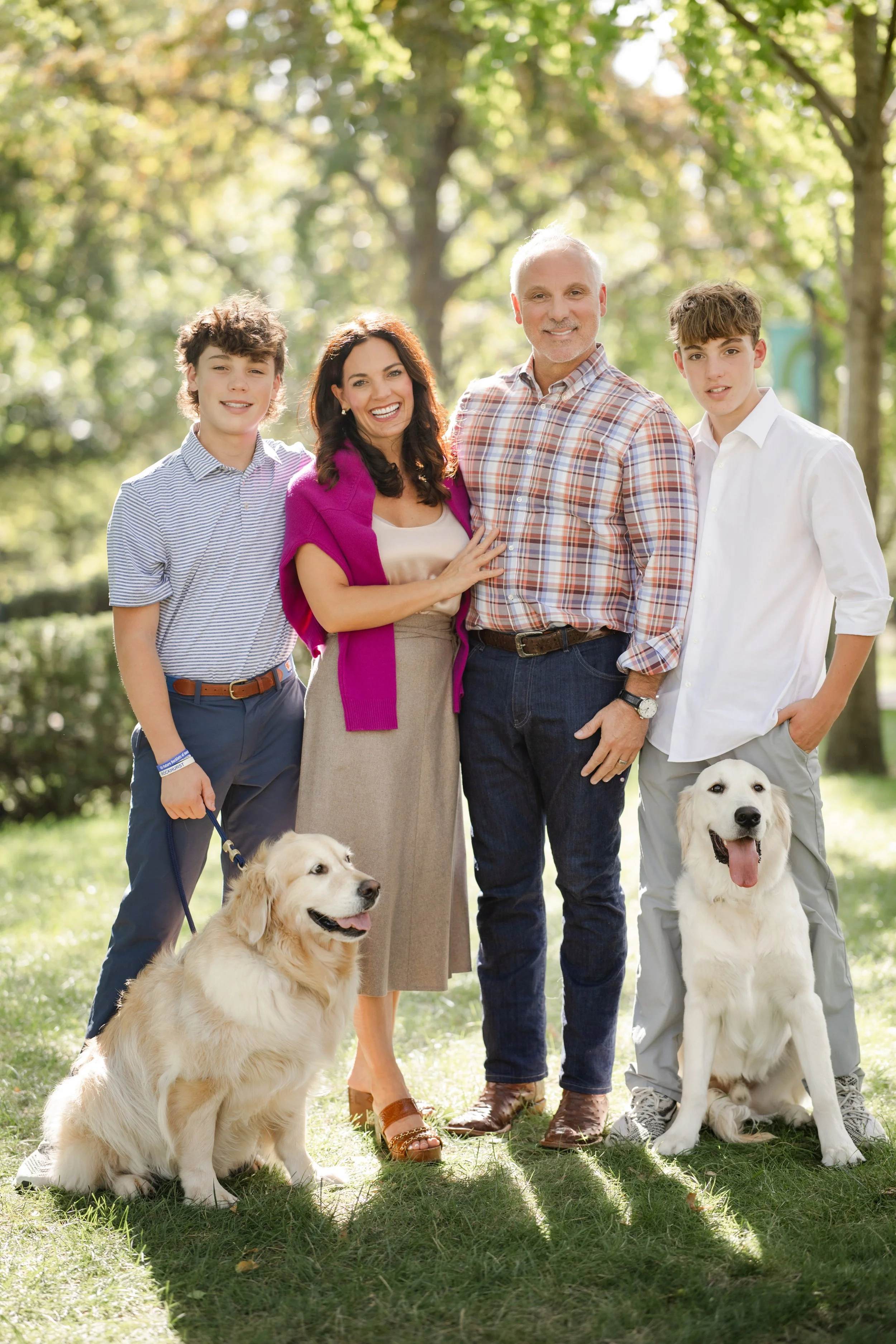 A happy family of four with two dogs standing outdoors in a park with trees. The family consists of a woman, a man, and two teenage boys. The woman is wearing a beige skirt and a purple jacket, the man is in a plaid shirt, and the boys are in dress shirts. The dogs are large, one with a golden retriever breed and the other a Labrador retriever, both sitting on the grass.