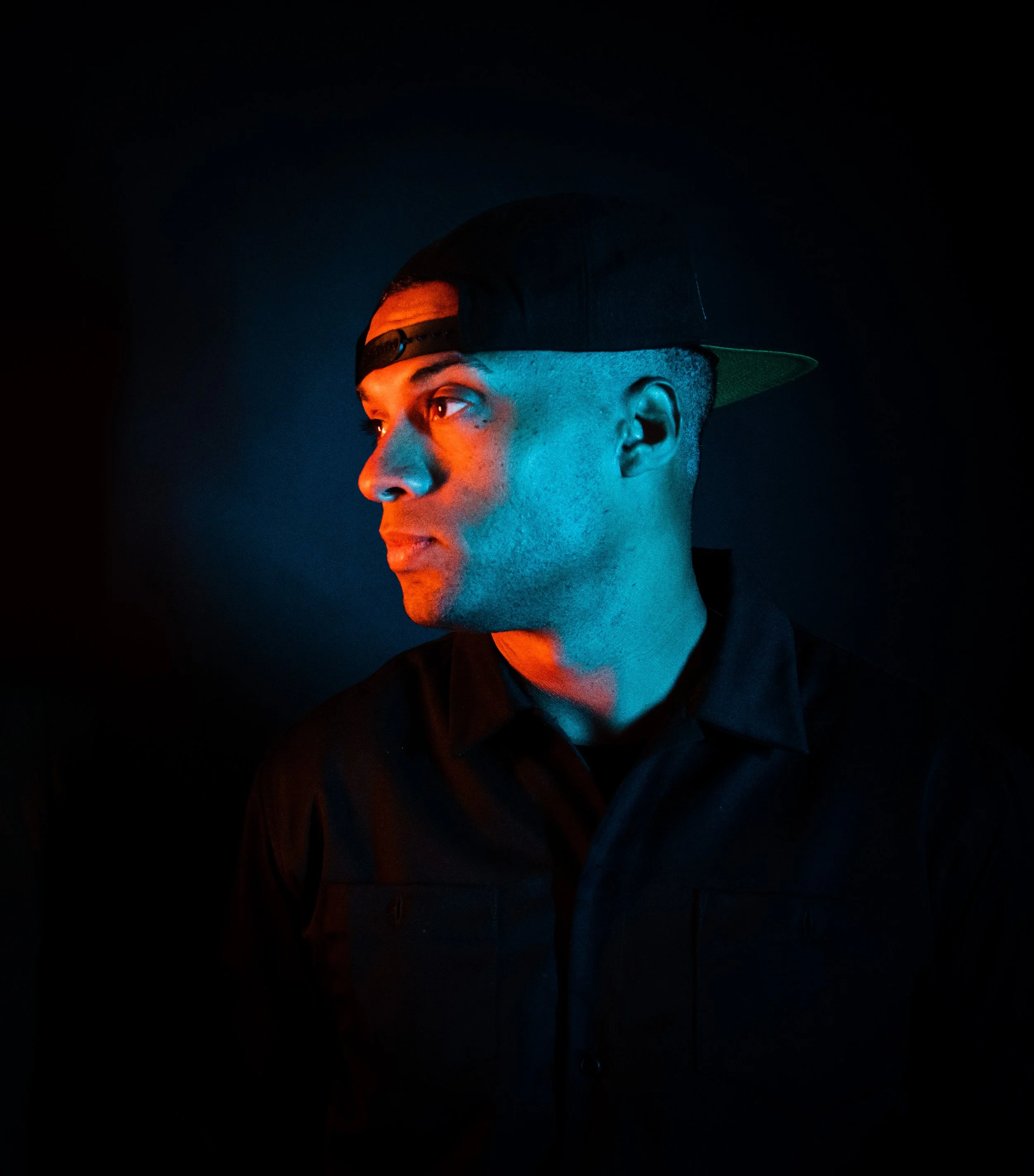 A young man, Dynamite MC,  wearing a black baseball cap backward with a flat brim and sunglasses, facing left, with blue and orange lighting creating a dramatic effect in a dark background.