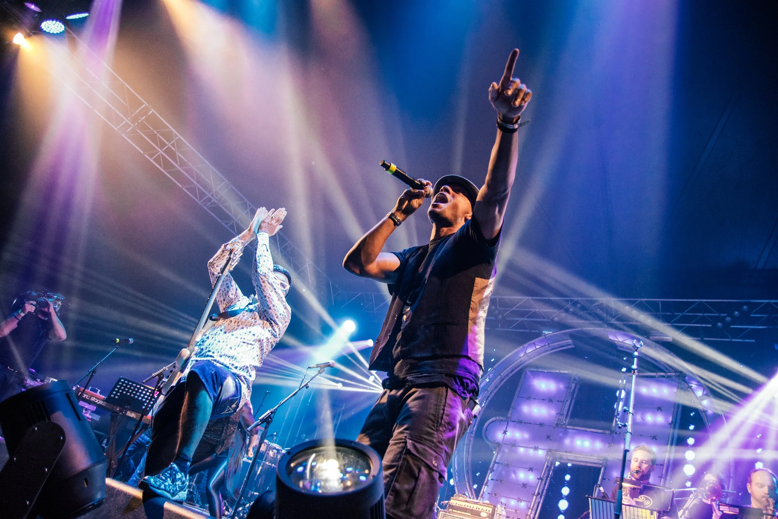 A lively concert scene with a singer, Dynamite MC  in a black shirt and hat, passionately singing into a microphone, and a guitarist with glasses in a patterned shirt, both illuminated by vibrant stage lighting and a large structured background.