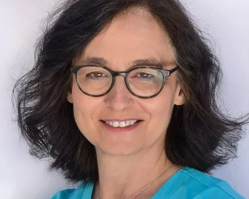 Portrait of a woman with dark, curly hair and glasses, smiling, wearing a blue top, against a plain background.