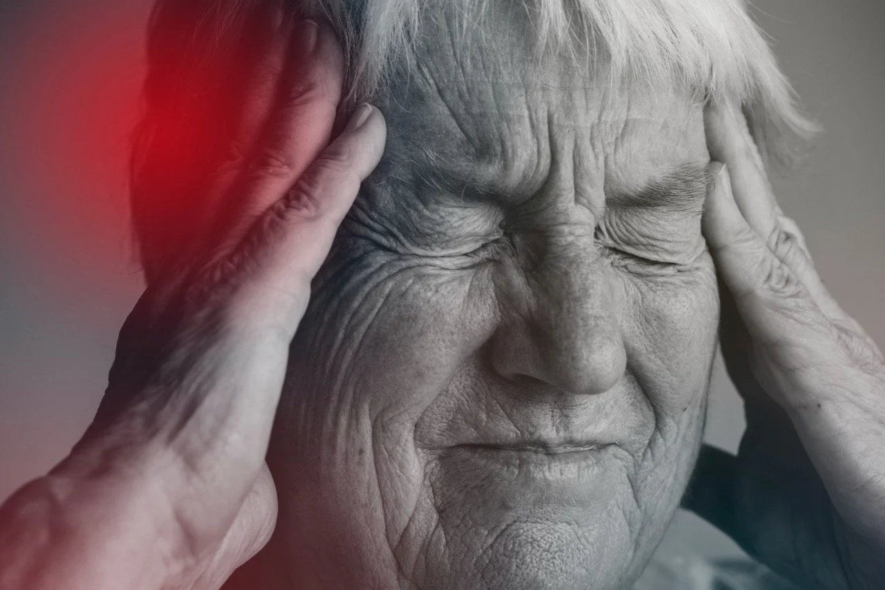 An elderly woman with closed eyes, holding her head with both hands, showing signs of pain or discomfort.