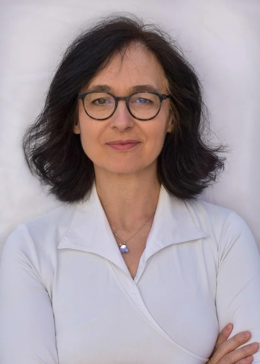 A woman with shoulder-length dark hair, glasses, and light skin, wearing a white shirt and a necklace, standing against a plain white background.