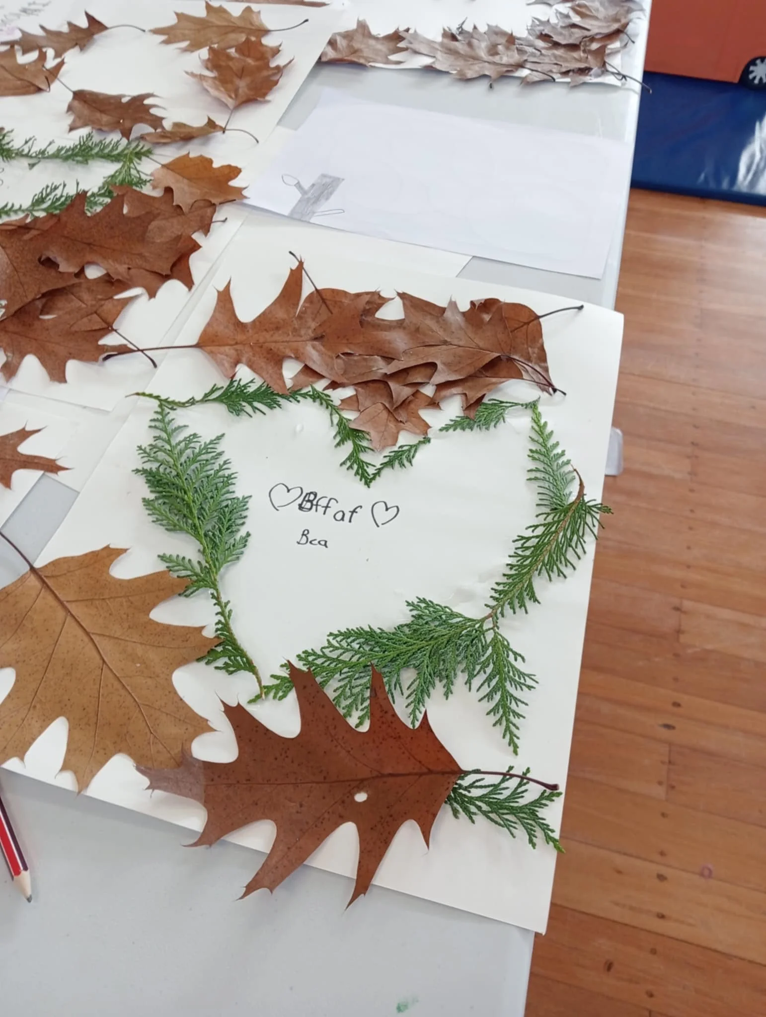 A table decorated with a variety of autumn leaves, including brown maple leaves and green fern leaves. There is a handwritten note on white paper that says '<3 Bf aft <3 Bea'. A pen is visible at the edge of the table, and the floor looks like wood.