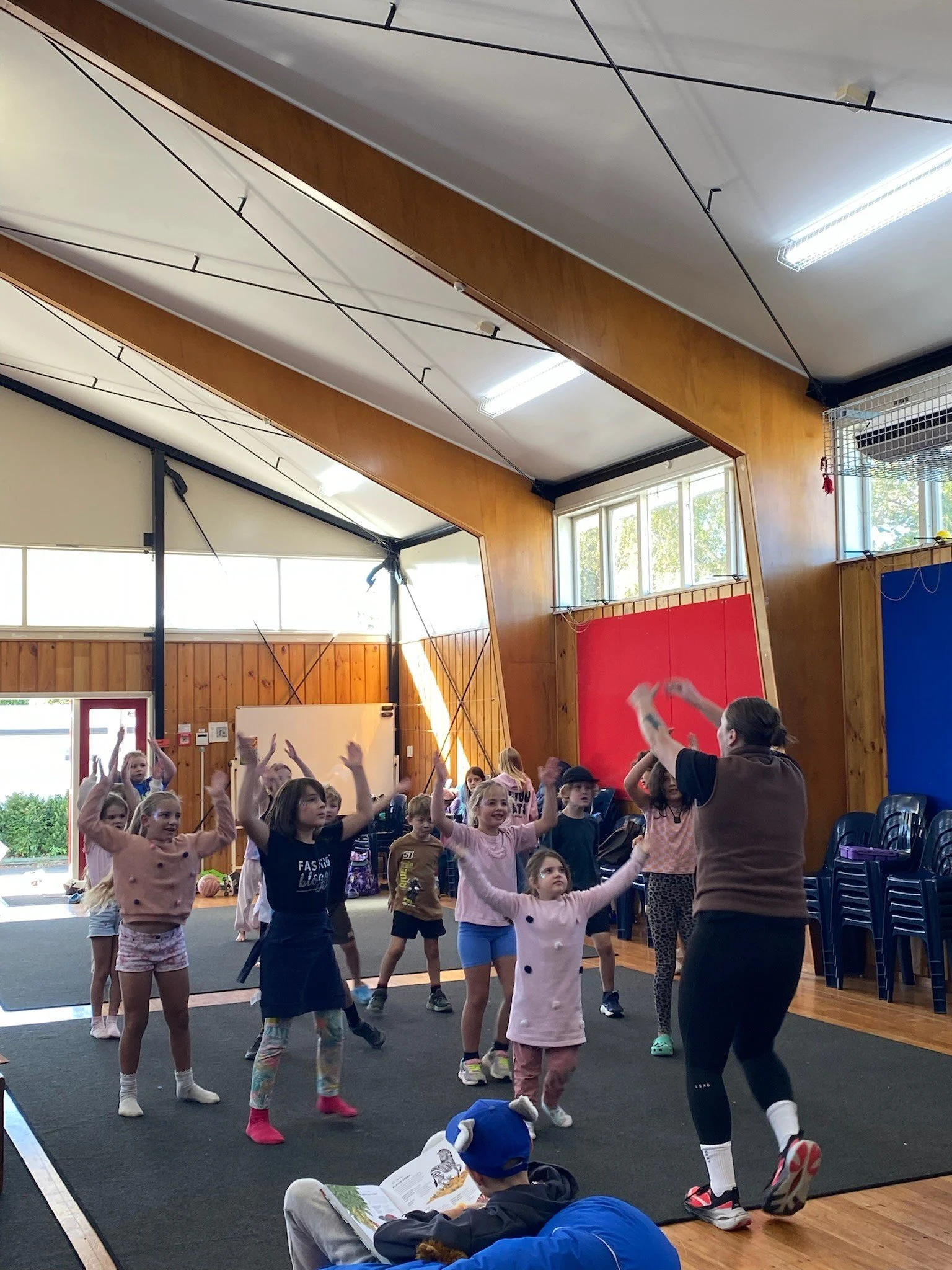 Children and an adult participating in a dance or exercise activity inside a gymnasium with wooden walls, large windows, and colorful gym mats.