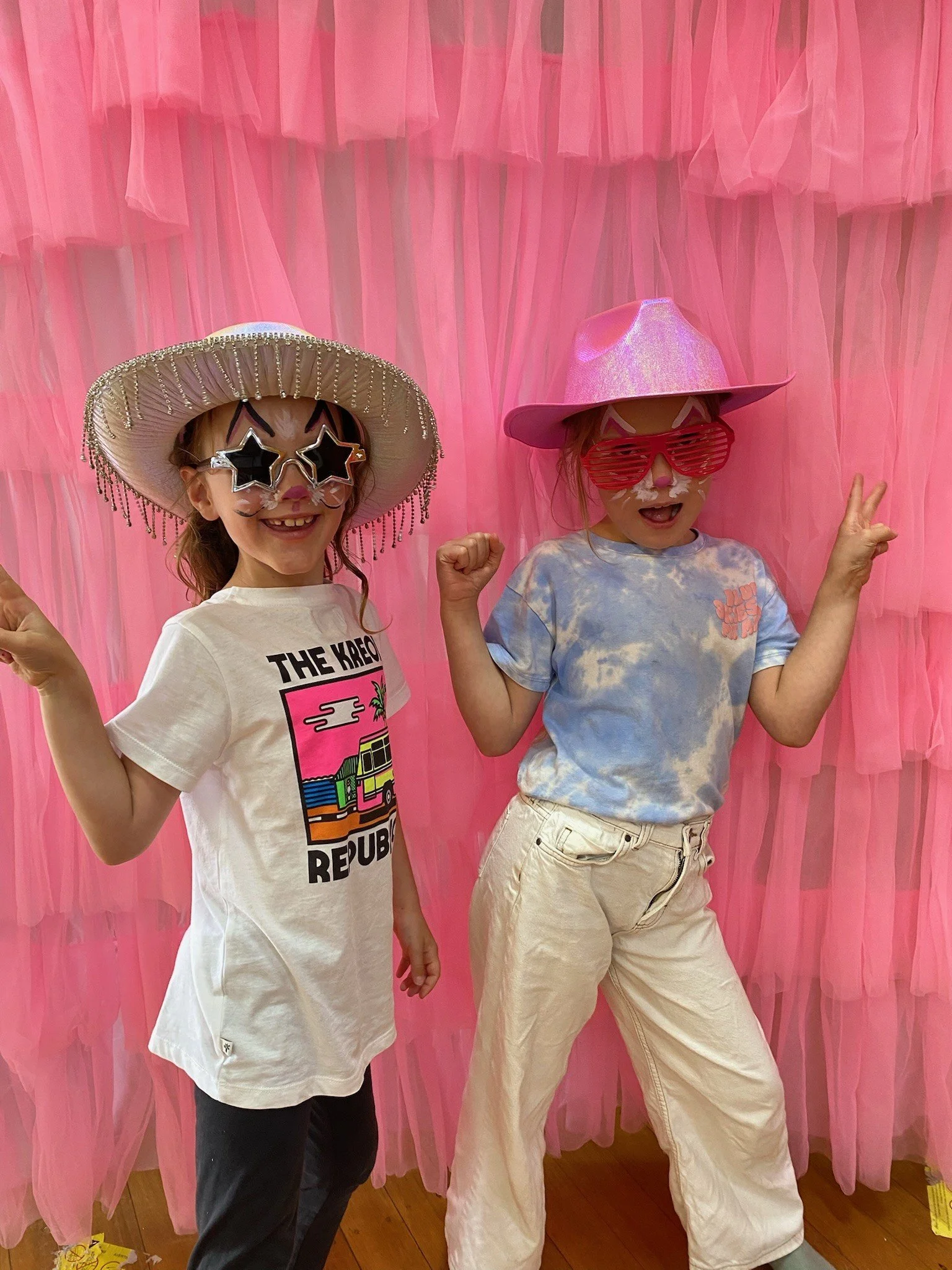 Two children posing in front of a pink tulle backdrop, wearing large hats and fun sunglasses, smiling and making playful gestures.