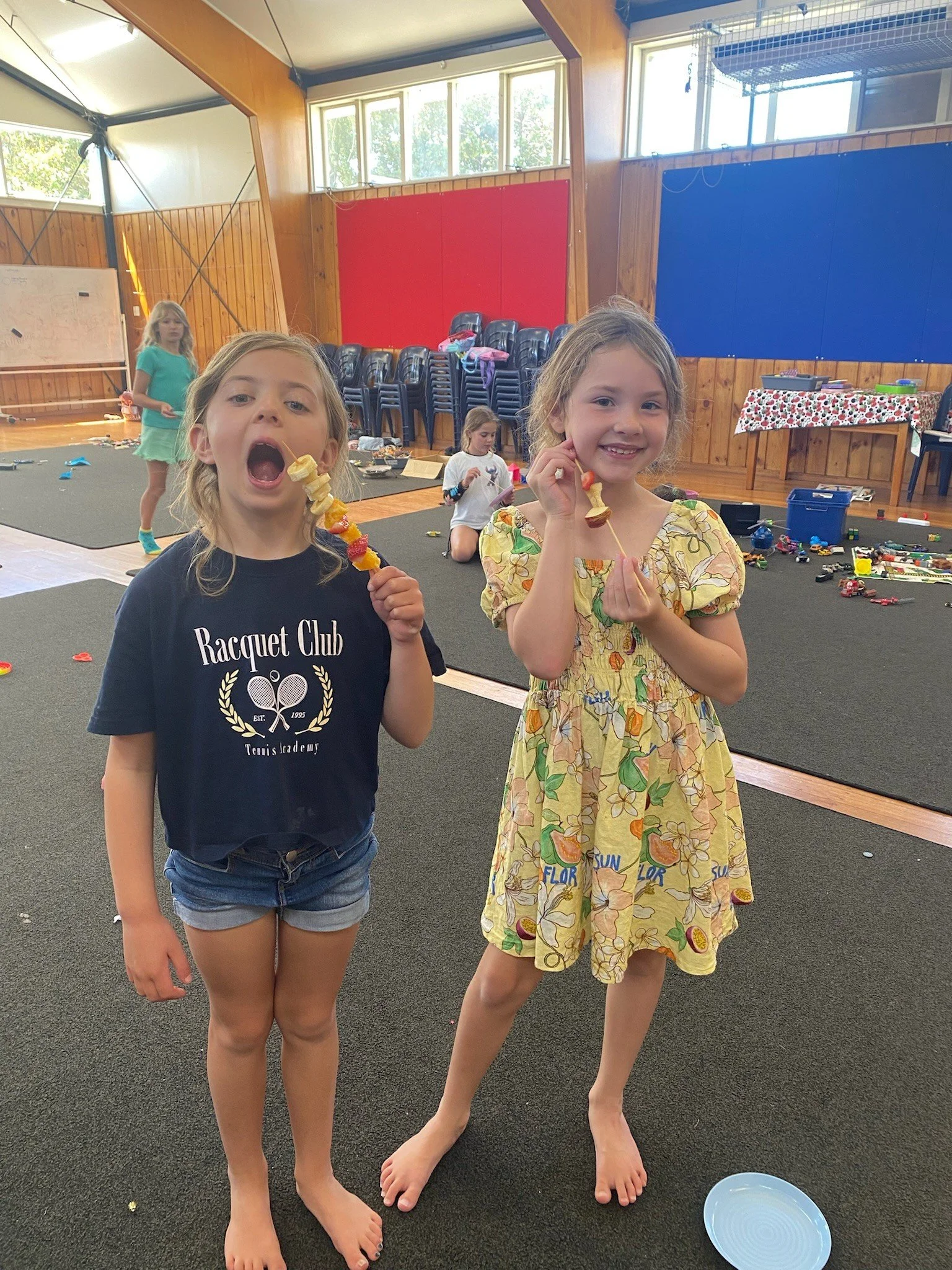 Two young girls standing indoors, holding skewers with fruit, smiling at the camera. In the background, other children are sitting and playing on the floor.