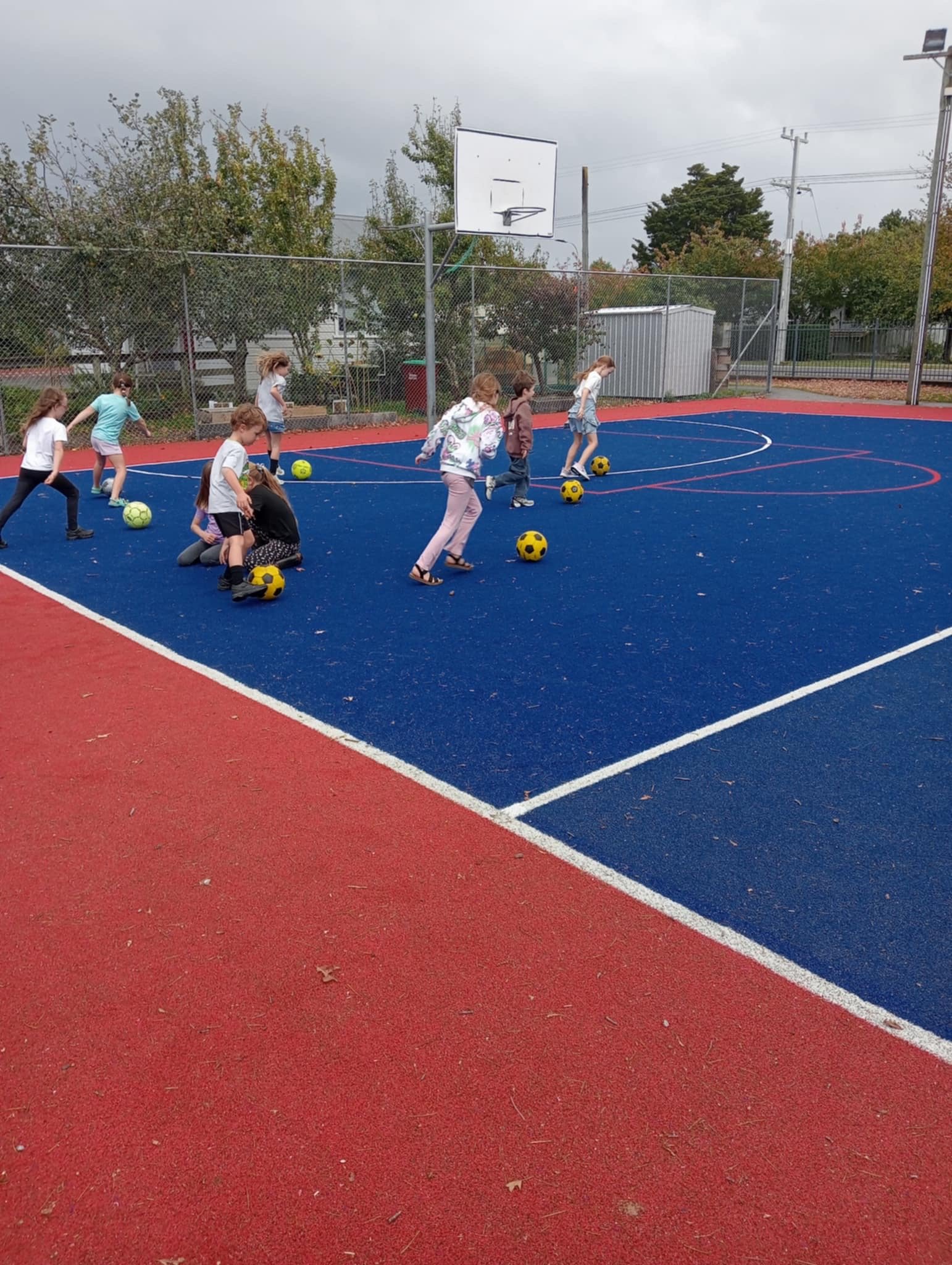 Children playing soccer on a blue and red outdoor basketball court, with a basketball hoop and chain-link fence in the background, trees, cloudy sky, and power lines.