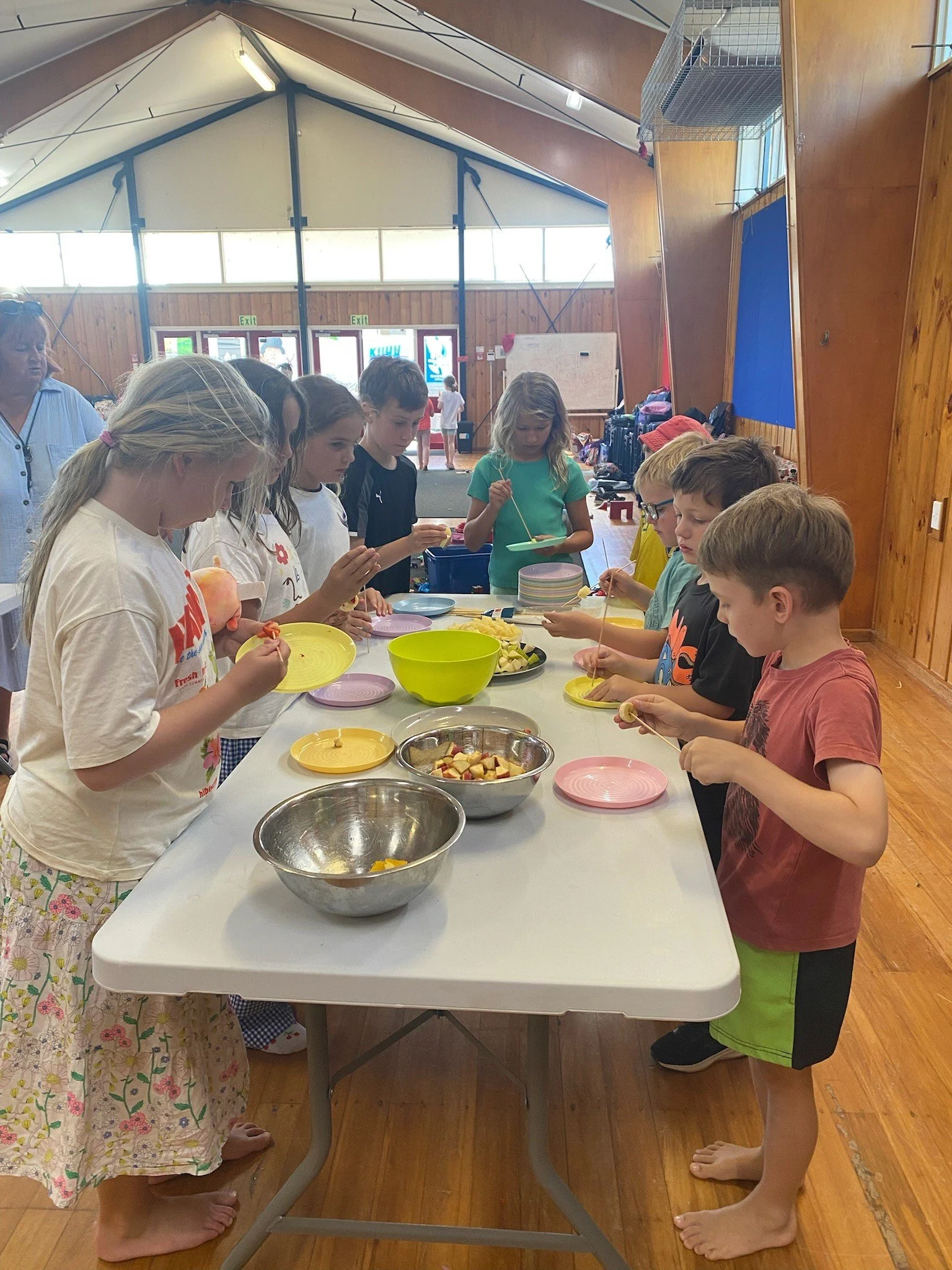 Children and an adult preparing food at a table in a community hall with wooden floors and walls, some children are concentrating on chopping or eating fruit, and bowls of prepared fruit are on the table.