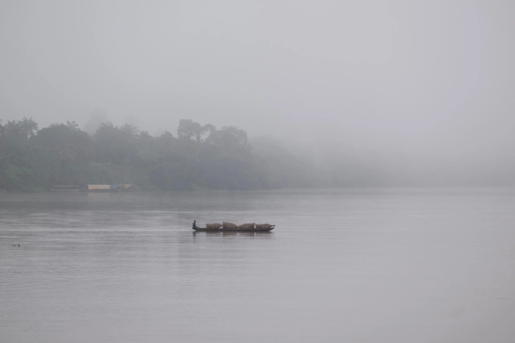 Niebla sobre un río con una pequeña embarcación y árboles al fondo.