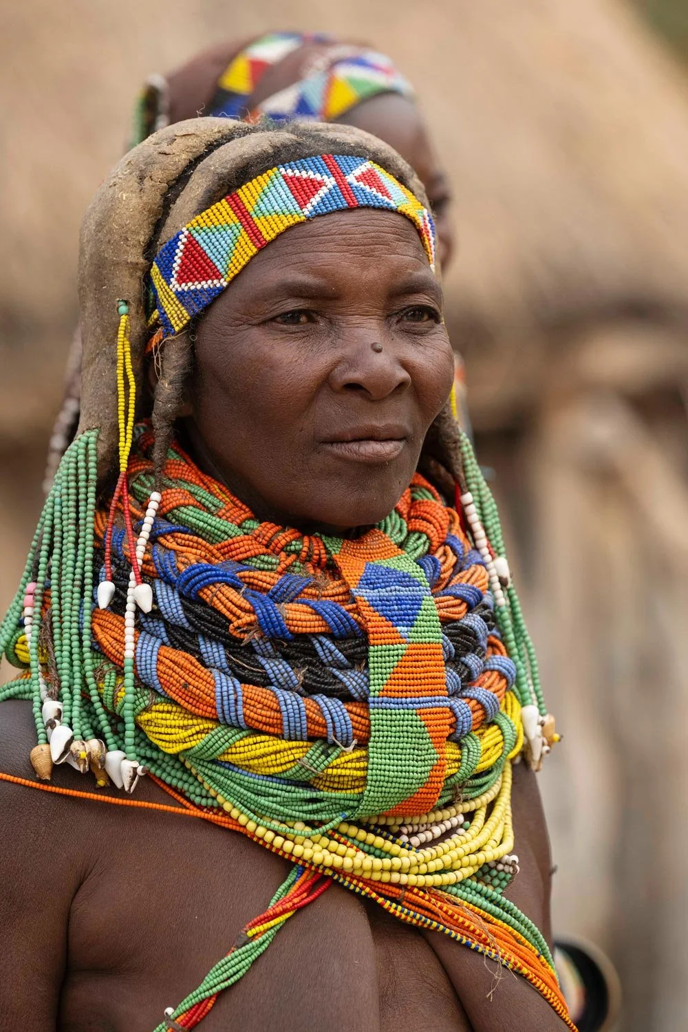 Retrato de una mujer indígena con accesorios y prendas coloridas, vestida con múltiples collares y un tocado de tela y cuentas, en un entorno natural.