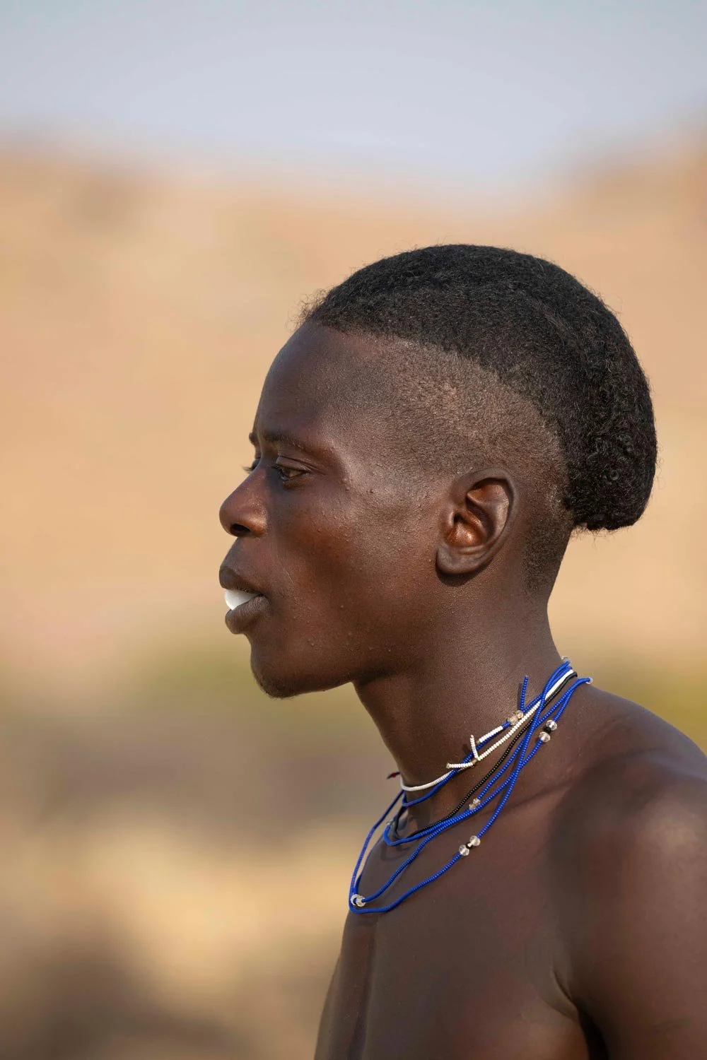 Retrato de perfil de un joven con piel oscura, cabello corto y peinado en forma de moño, usando varias cadenas de collar en color azul y blanco, en un fondo de paisaje desértico.