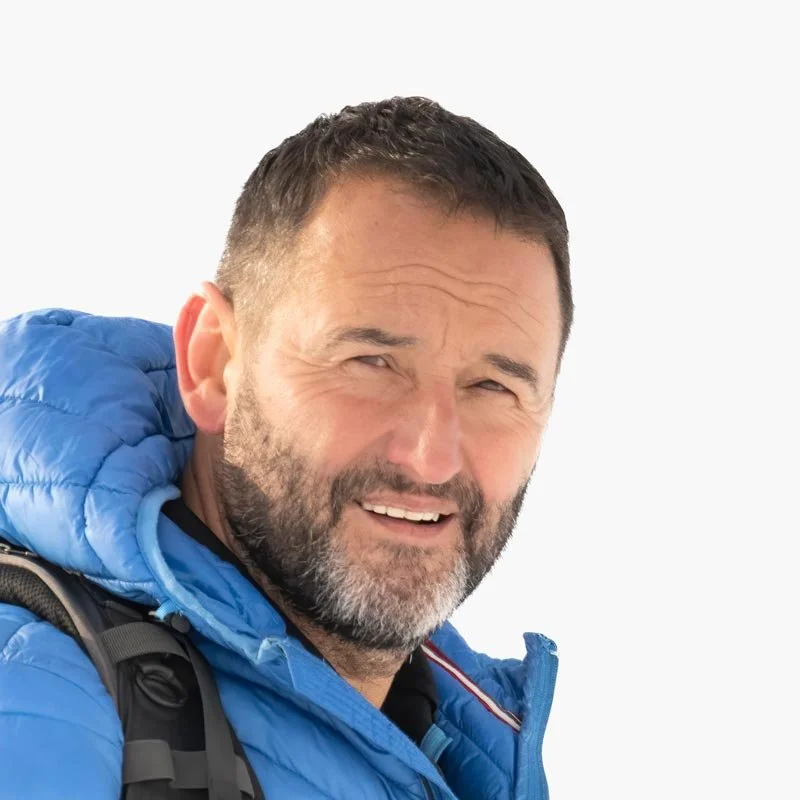 Hombre con barba, usando chaqueta azul y mochila, sonriendo, fondo blanco.