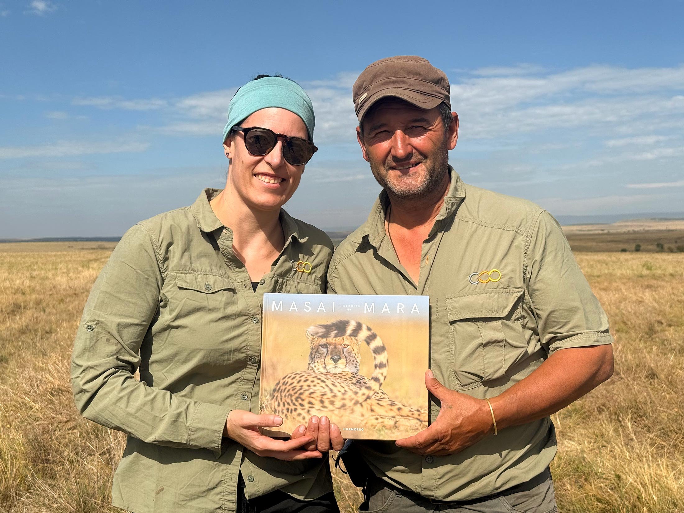 Dos personas sosteniendo una revista con una imagen de un guepardo en un paisaje de sabana. Ambos llevan camisas similares de color verde y parecen estar en una área de conservación o safari.