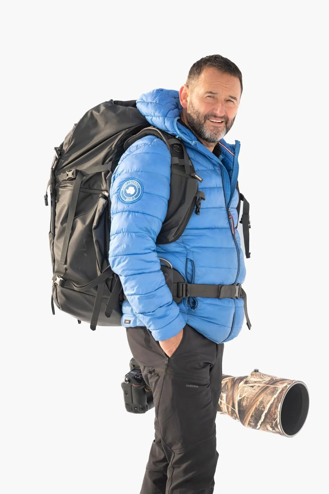 Hombre con ropa de montaña, mochila y cámara, sosteniendo un largo tubo de telescopio, en un fondo blanco.