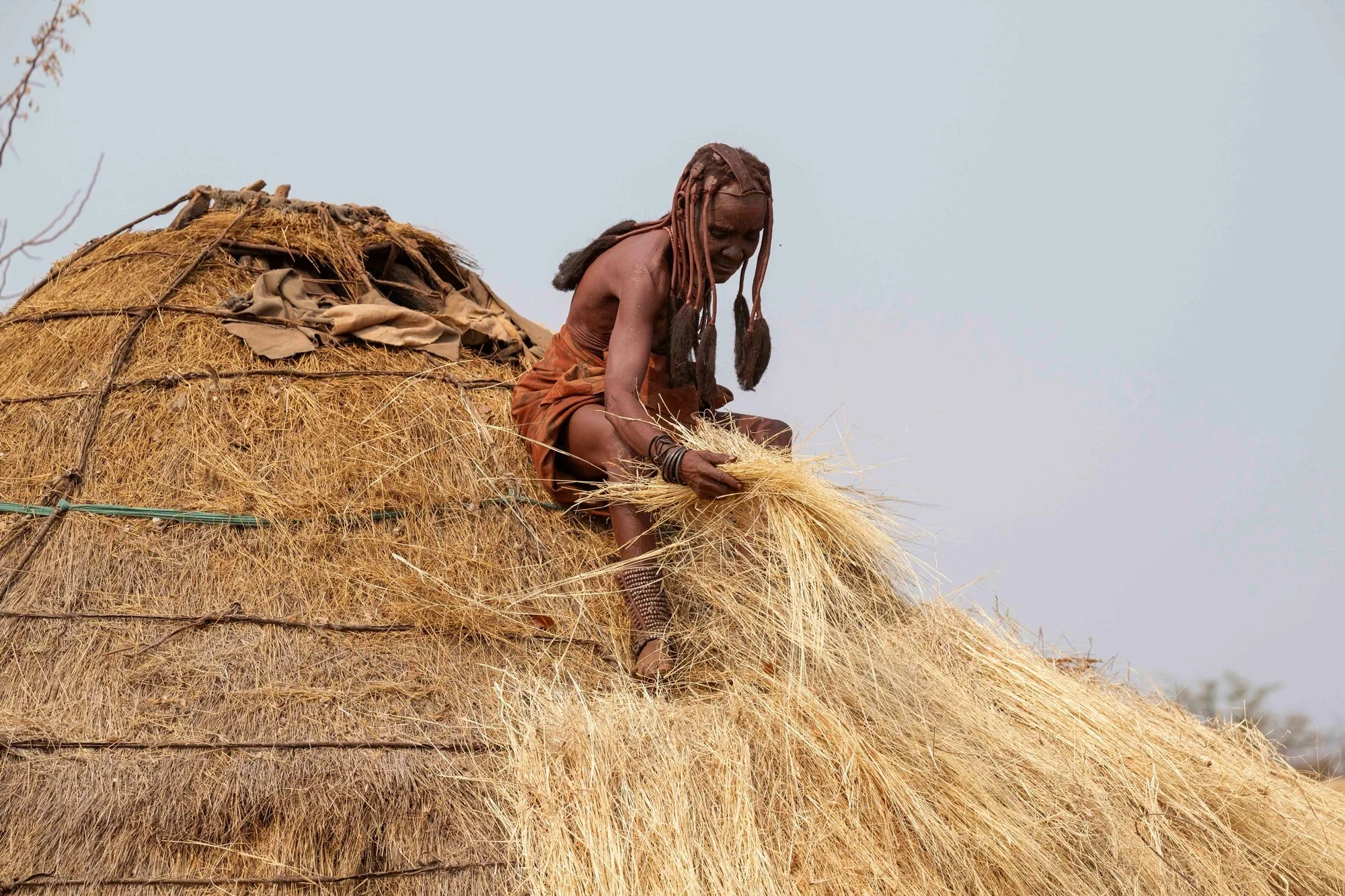 Anciano africano con rastas, sentado sobre un gran montón de paja, atándose un mechón de paja, con fondo de cielo despejado.