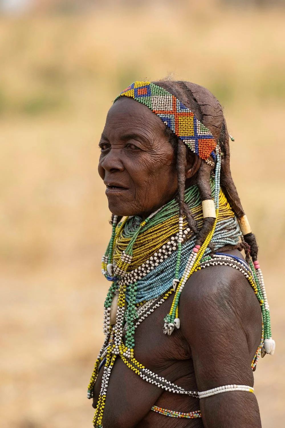 Mujer de África vestida con múltiples collares y accesorios tejidos con cuentas, usando una banda colorida en la cabeza y su cabello en rastas.