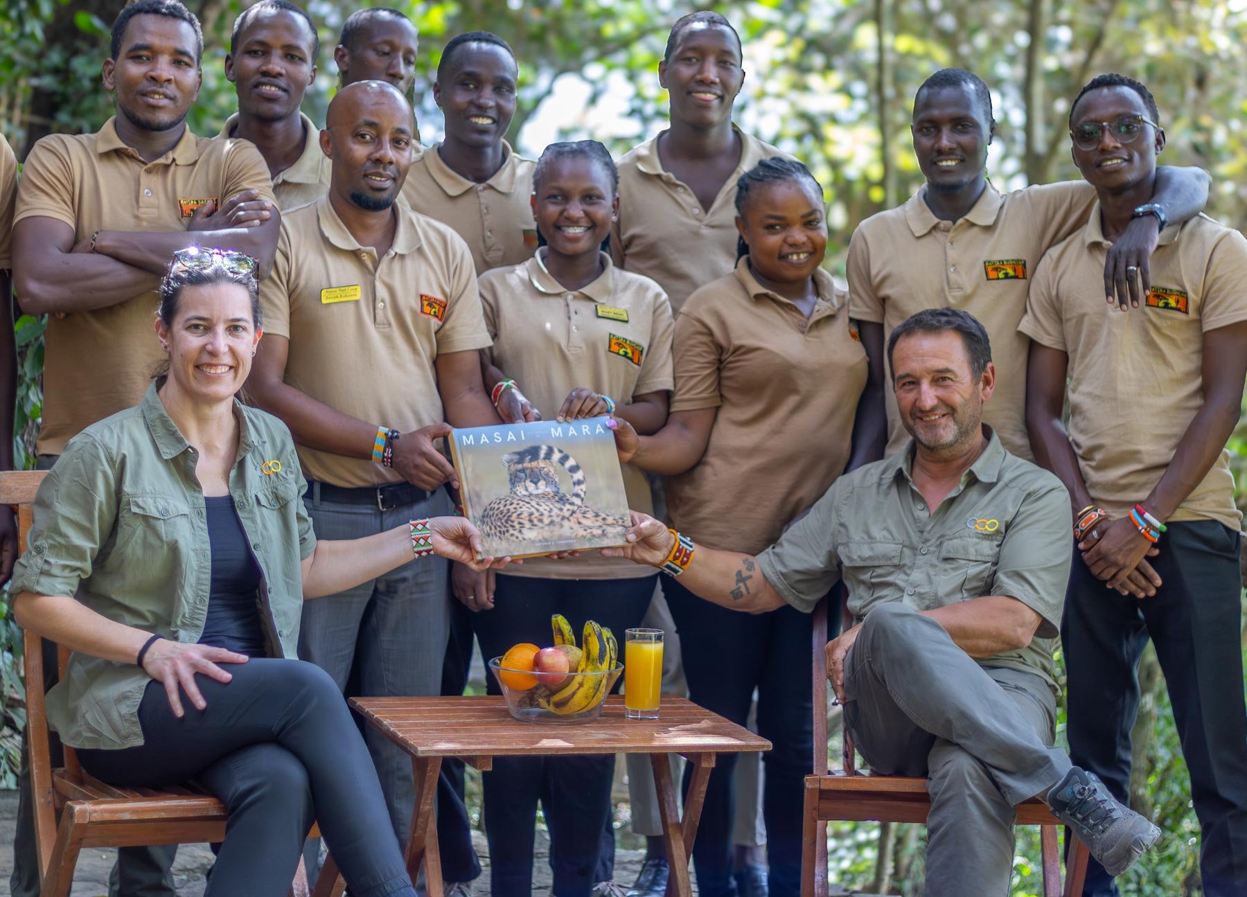 Grupo de personas en un entorno natural, algunos con camisetas beige con logotipos, sosteniendo un libro con la imagen de un guepardo y el título 'Masai Mara'. En una mesa, hay fruta y jugo de naranja. Dos personas están sentadas y las demás de pie.