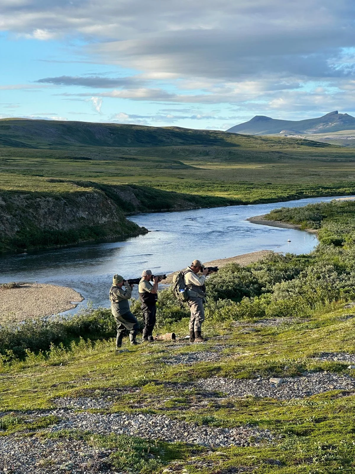 Tres personas con equipo de explorador observan un río en un paisaje de montañas y praderas.
