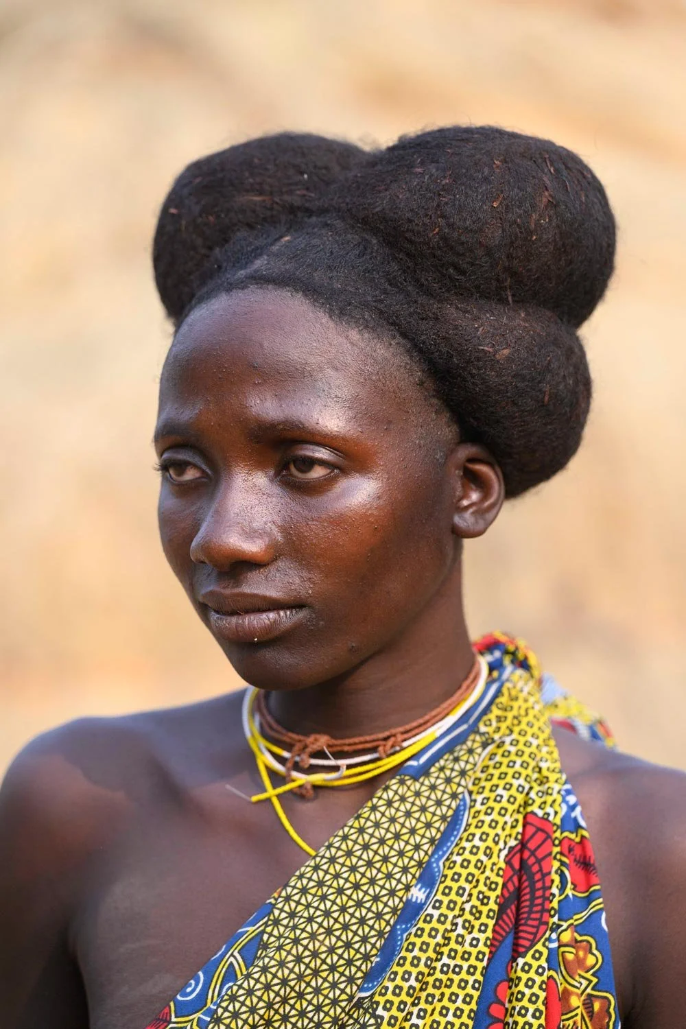 Retrato de una mujer con piel oscura, cabello trenzado en estilo tradicional, vestida con tela colorida y collar de varias vueltas, en un fondo difuminado de color beige.