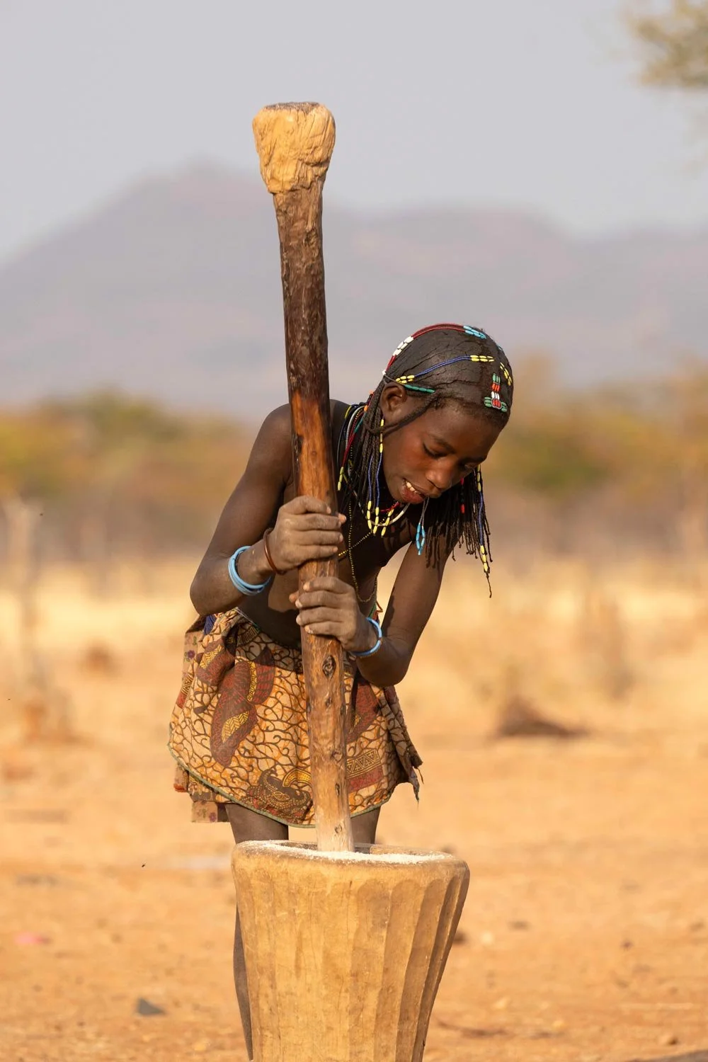 Mujer artesanal africana moliendo grano en un mortero de madera en un campo seco y árido, con montañas en el fondo.