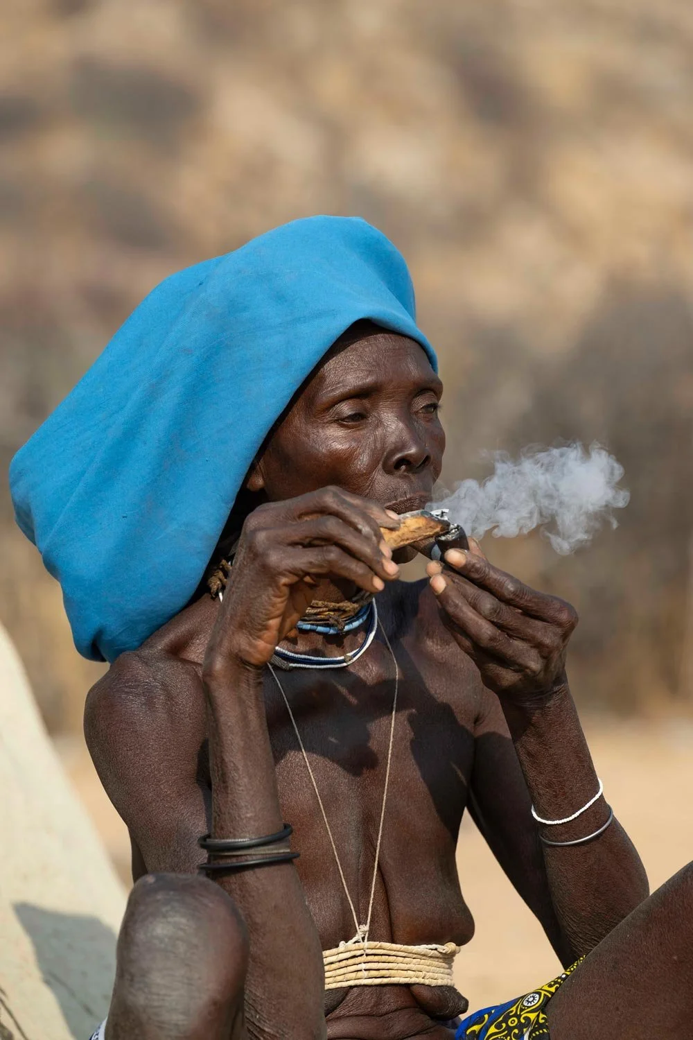 Anciano de piel oscura con un tocado azul, sentado y fumando. Lleva varias pulseras y collares, en un entorno natural árido.