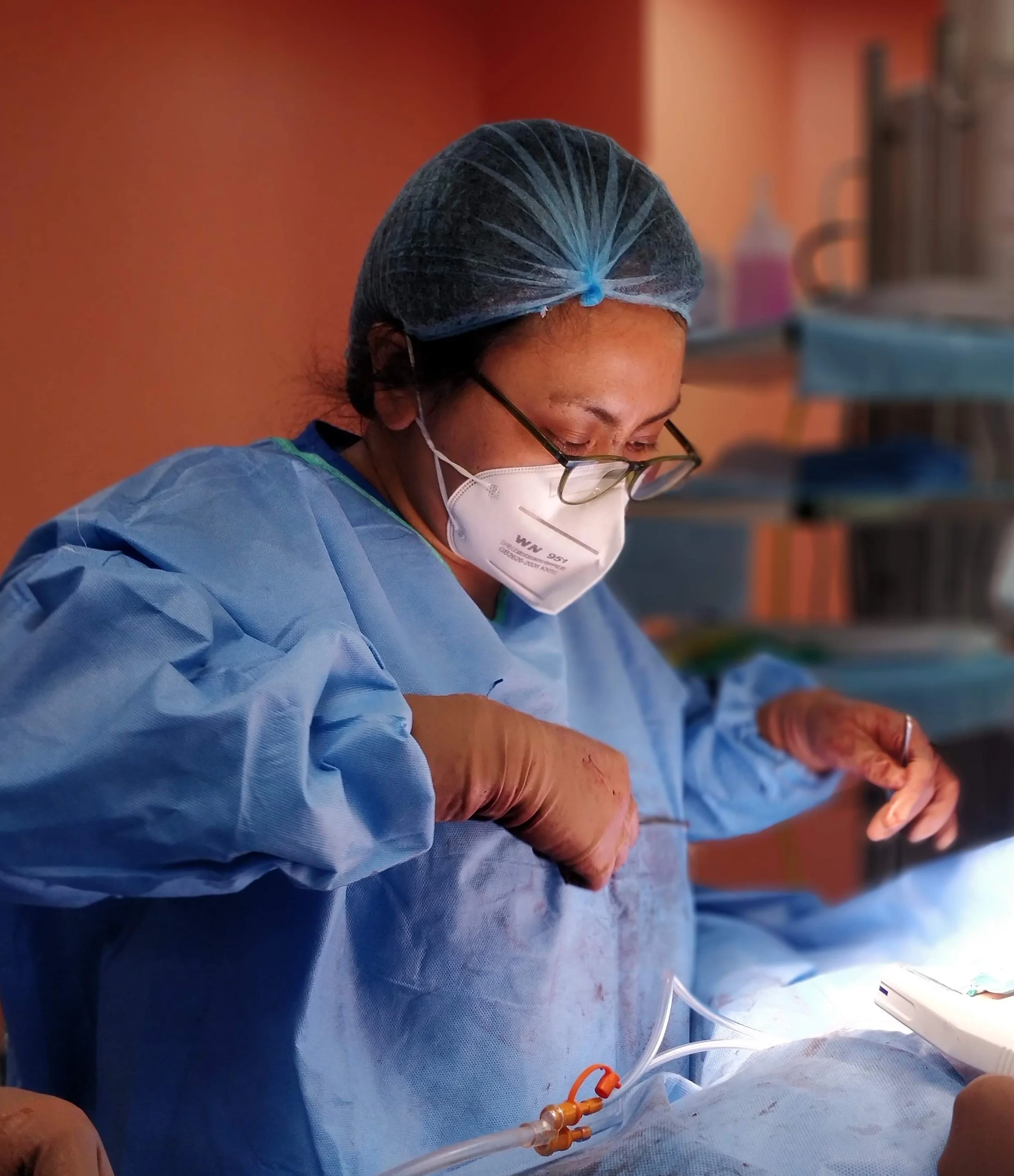 Dr Ananya Deori in surgical attire, including a hair cap, mask, and gloves, performing a procedure in an operating room with medical equipment and supplies in the background.