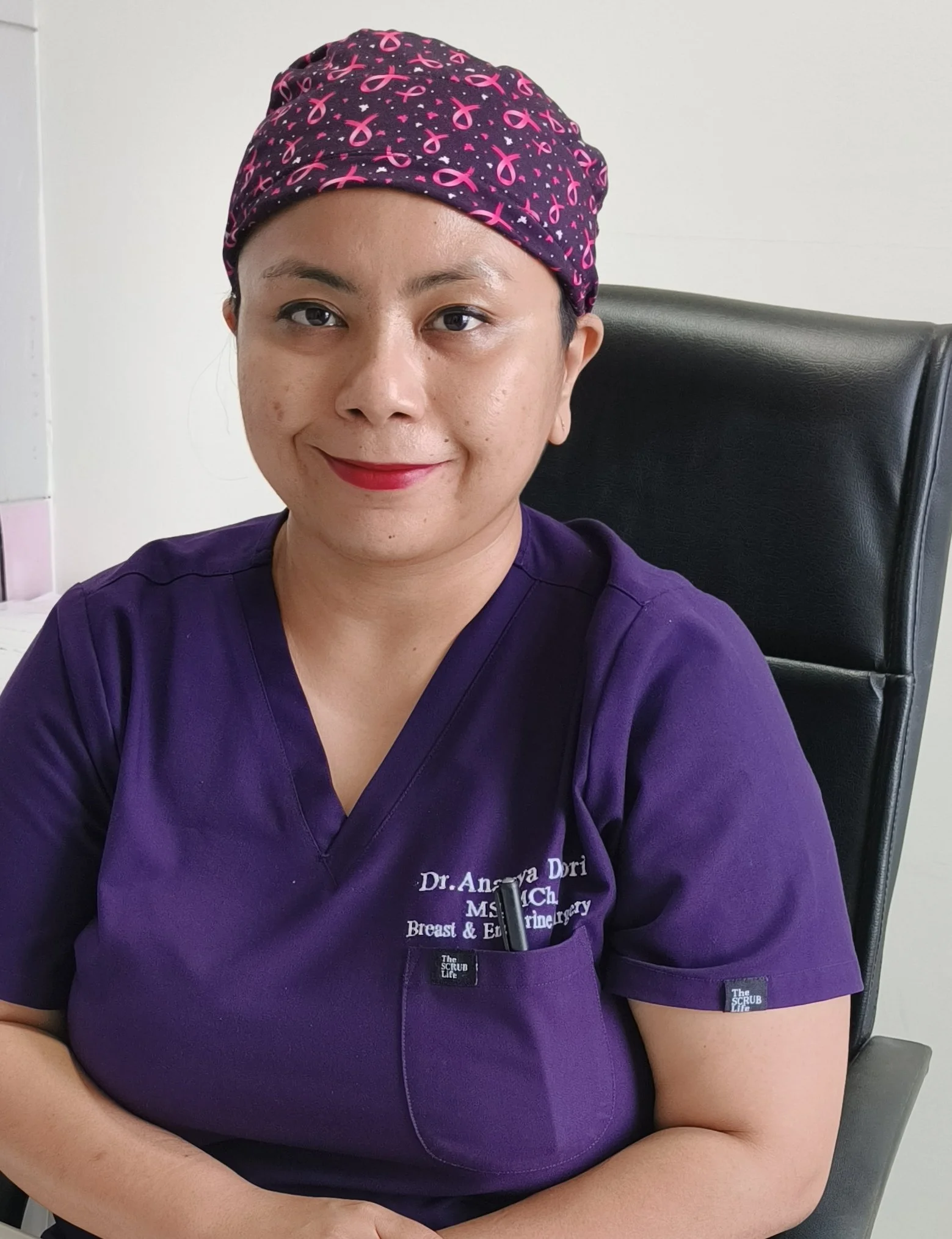 Close-up of a woman wearing maroon scrubs with embroidered text and a purple surgical cap with pink ribbons, sitting in a black chair, smiling.