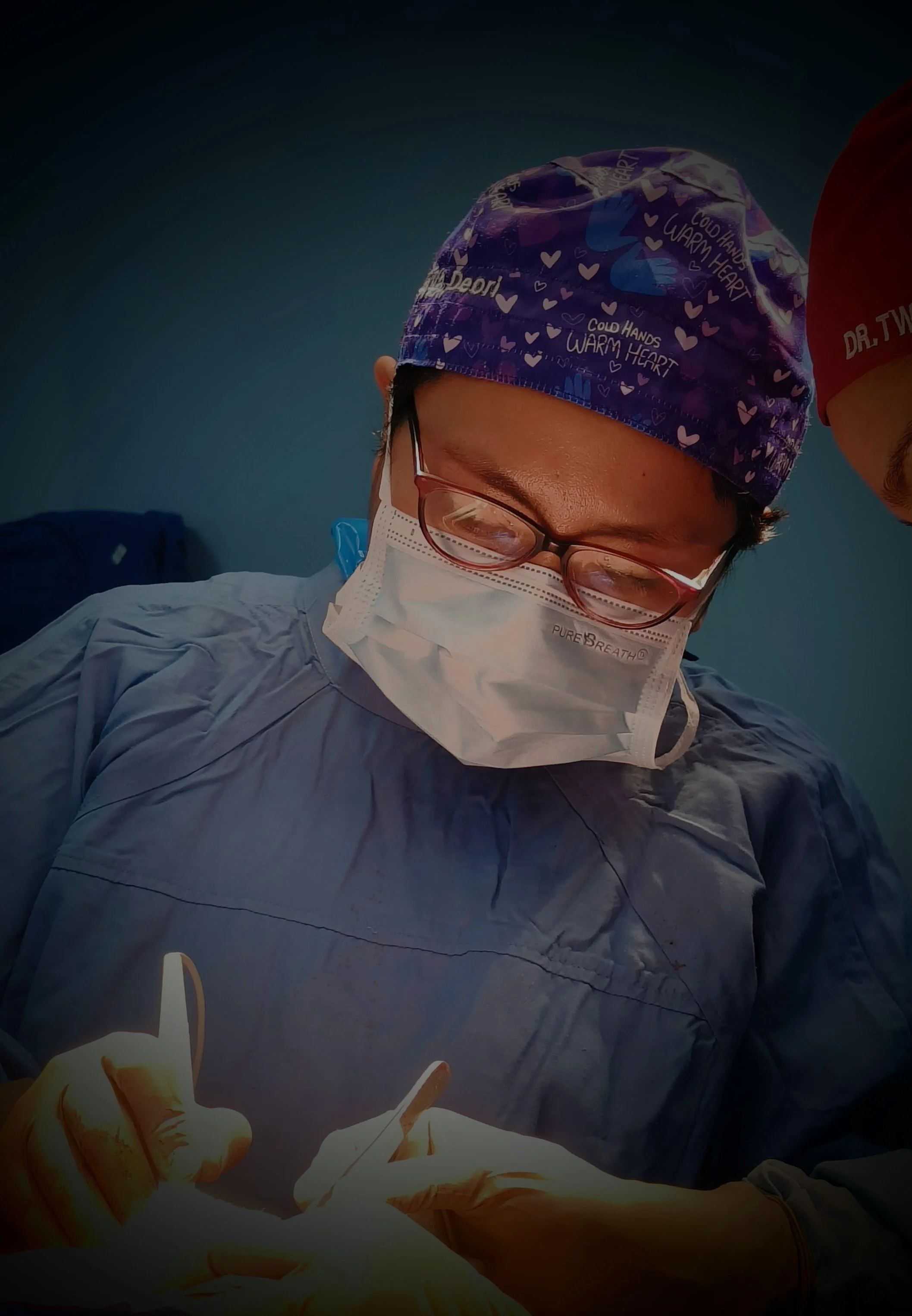 A surgeon wearing a purple cap with hearts and phrases, a face mask, glasses, and surgical scrubs performs an operation in an operating room.