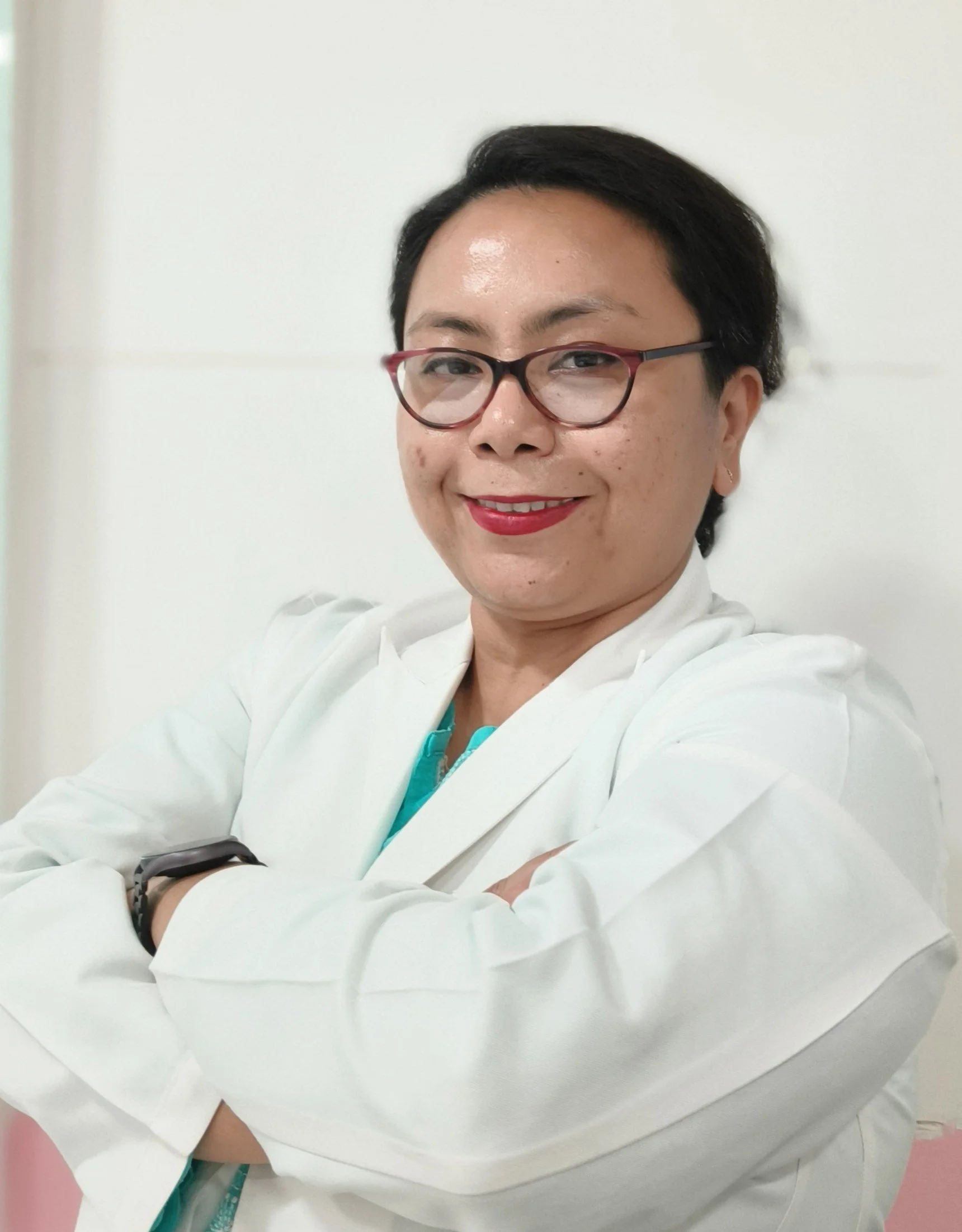 A woman in a white lab coat smiling, wearing glasses, with arms crossed, standing against a plain light-colored wall.