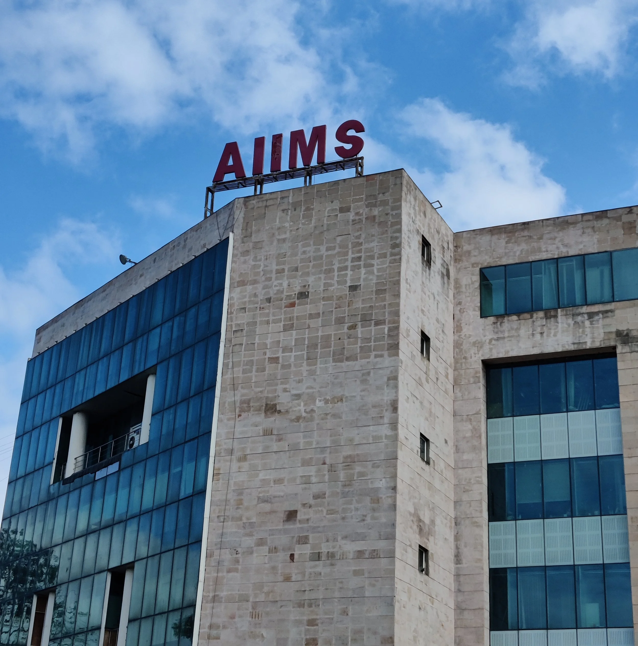 A modern multi-story building with a stone facade and blue-tinted glass windows, topped with a red sign that reads "AIMS" under a partly cloudy sky.