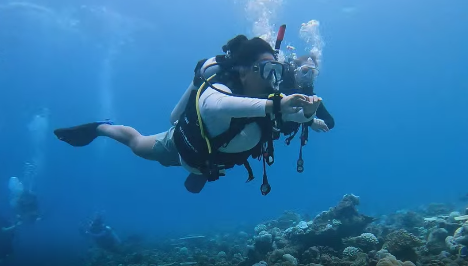 Person scuba diving underwater near coral reef, holding hand in front.