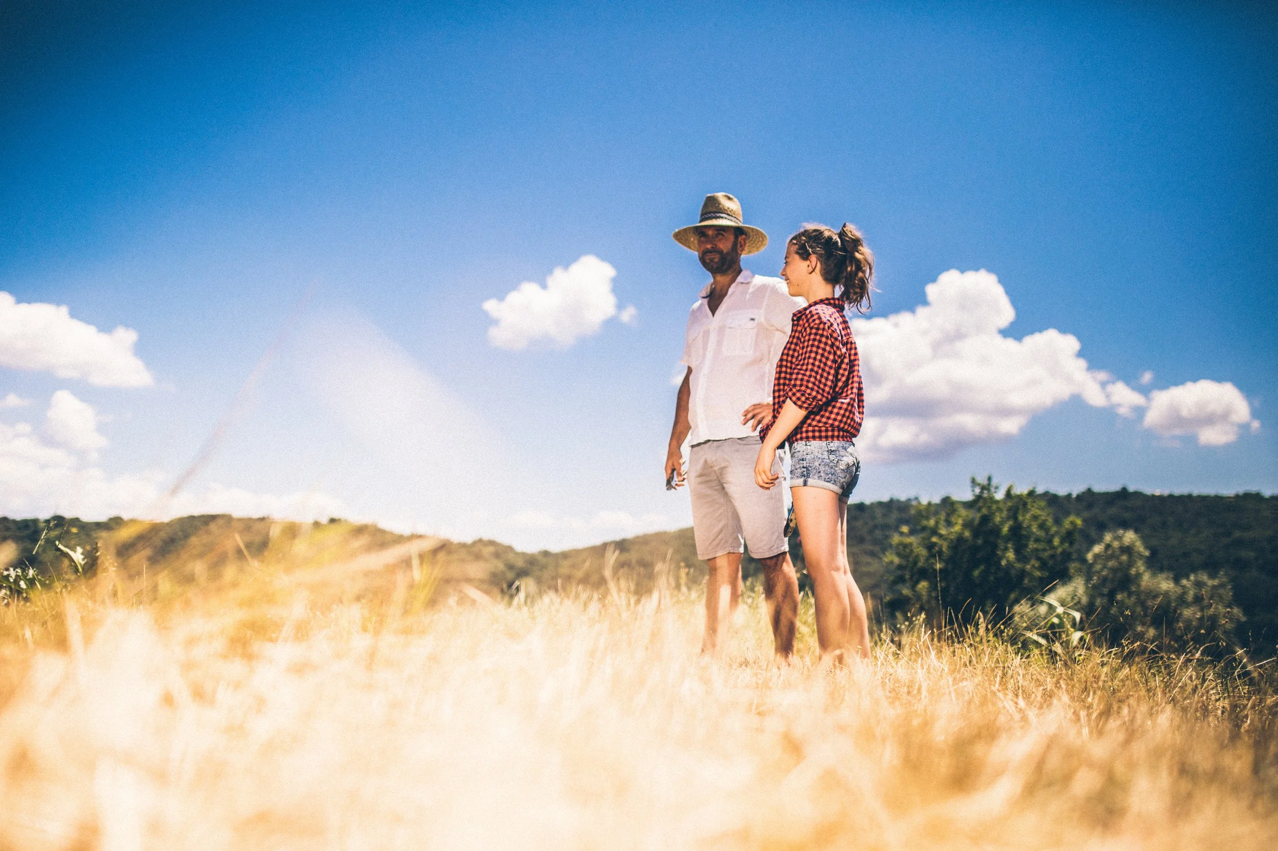 A man and a woman standing in a grassy field under a blue sky with scattered clouds, with mountains or hills in the background.