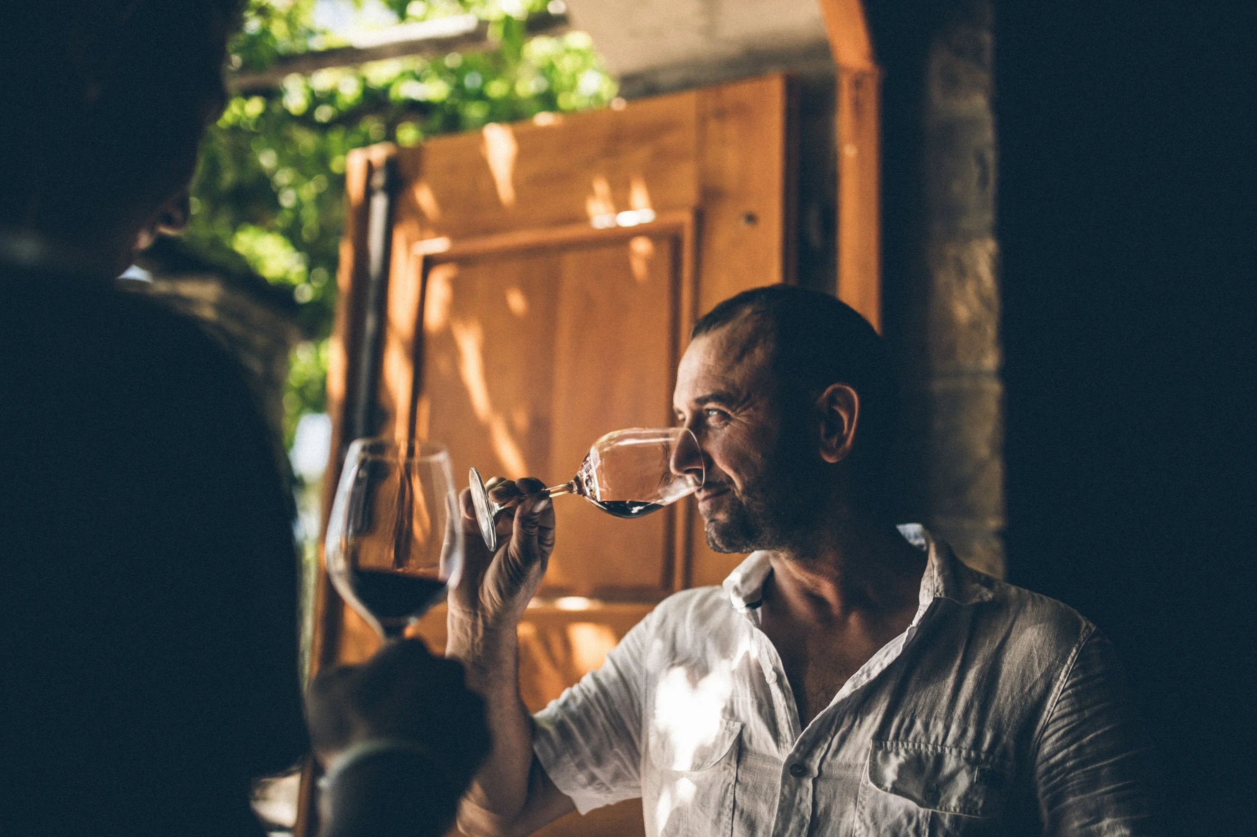 A man with short dark hair and a beard wearing a white shirt, sitting indoors, enjoying a glass of red wine, with another person partly visible on the left in a dimly lit setting with natural light and green foliage outside.
